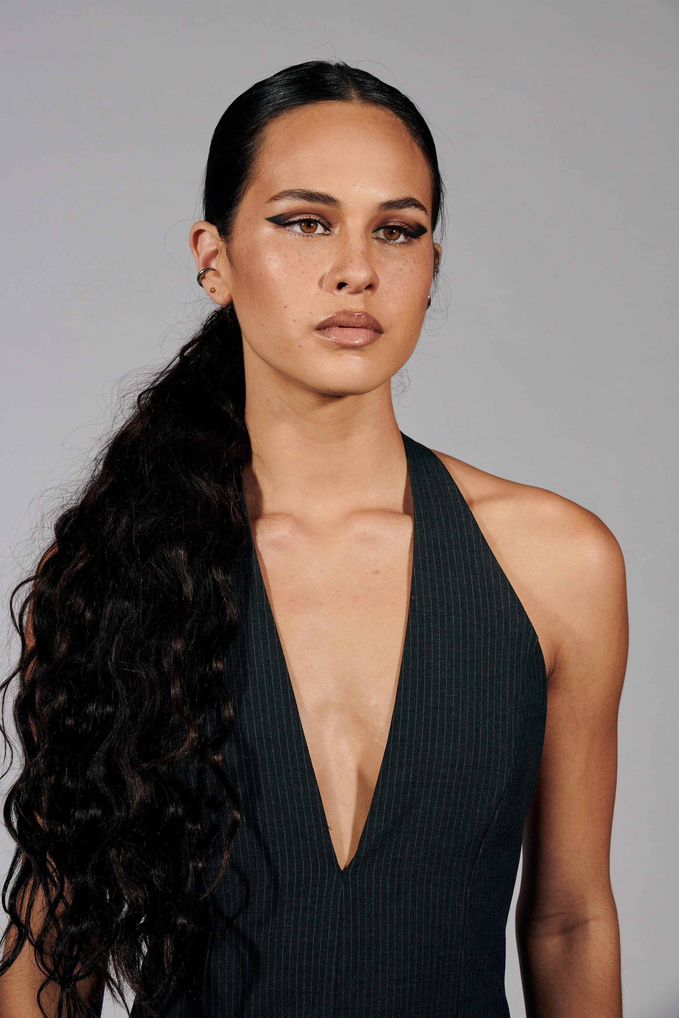 A woman with long, wavy black hair wearing a black, sleeveless top with a deep V-neck, looking directly at the camera with neutral expression against a plain gray background.