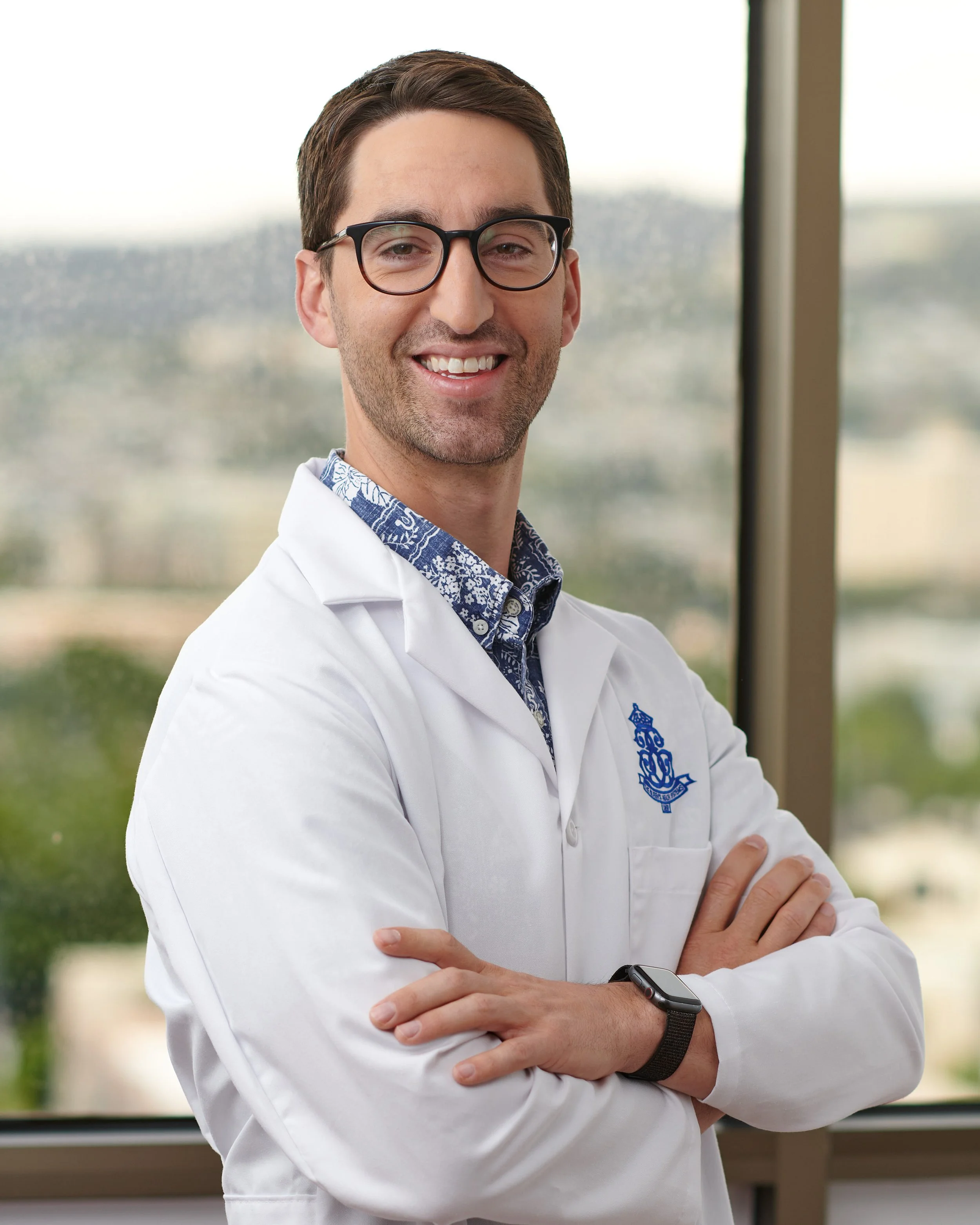 A smiling male doctor in a white coat with a blue emblem, glasses, and a smartwatch standing with arms crossed in front of a window.