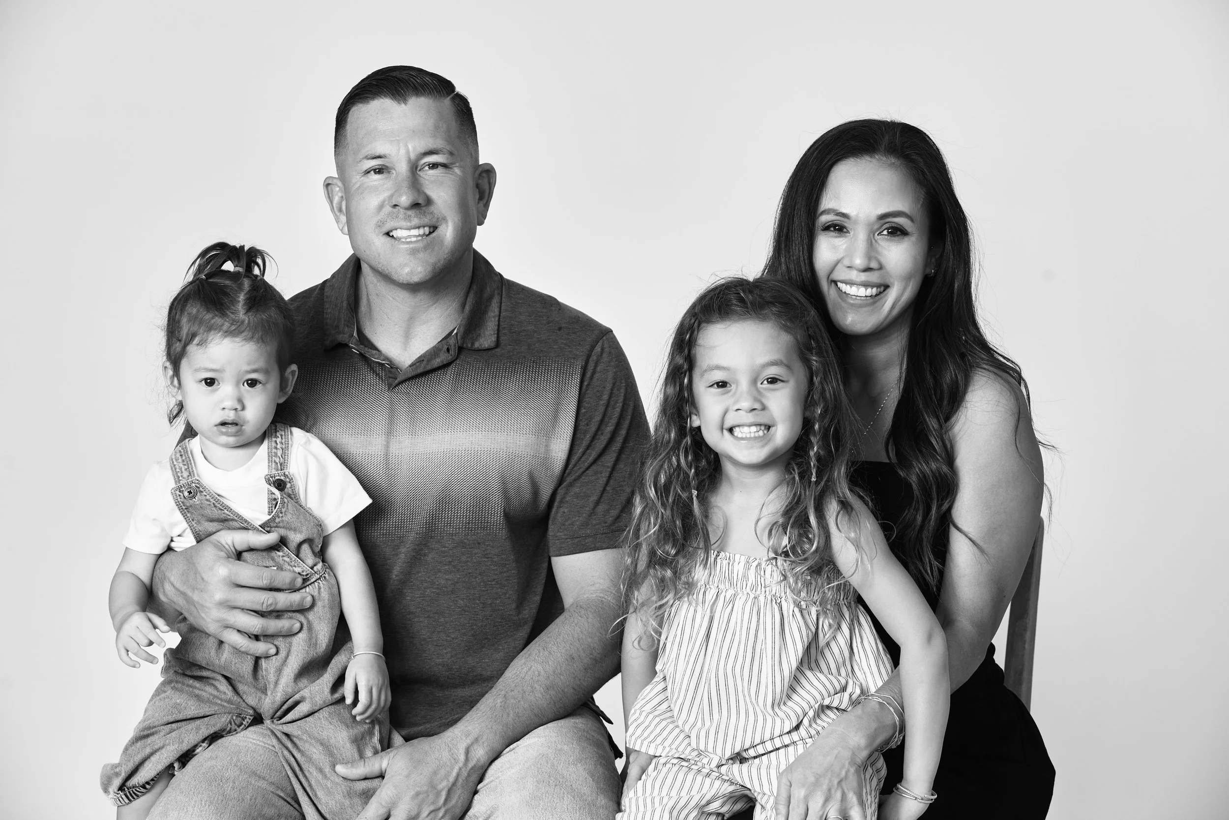 A black-and-white portrait of a multiracial family of five sitting together, smiling at the camera against a plain background. The family includes two adult women, a man, and two young girls.