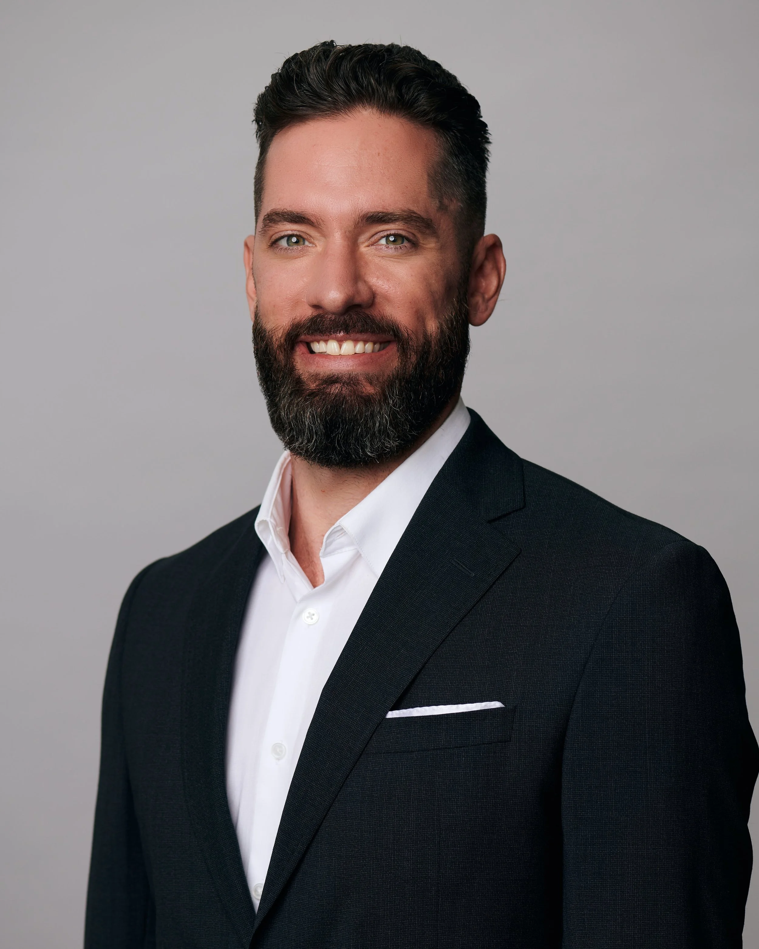 Headshot of a man with dark hair, a full beard, wearing a black suit and white shirt, smiling against a gray background.