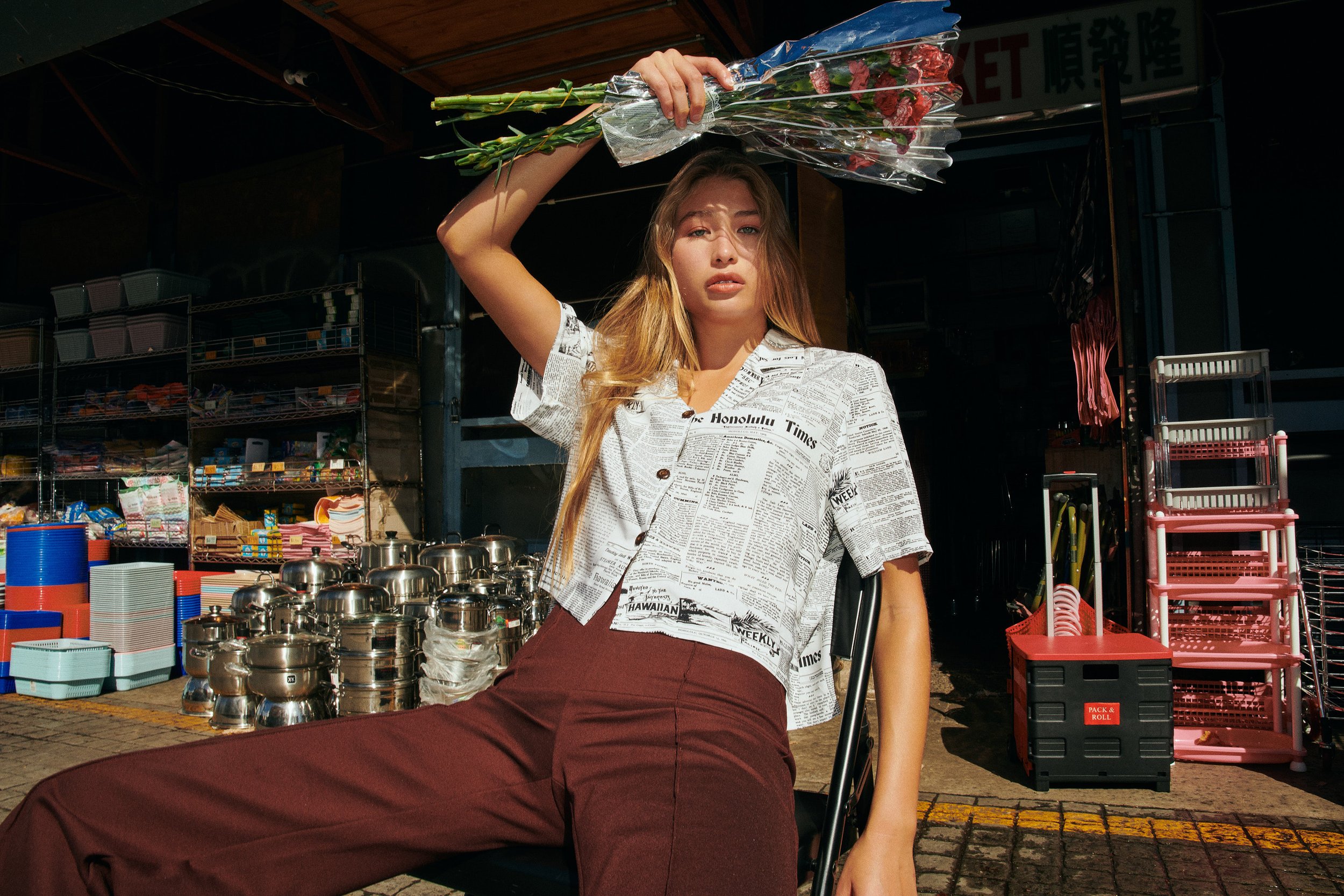A young woman with long light brown hair holds a bouquet of pink roses above her head while sitting on a black chair outside a market stall. She wears a short-sleeved shirt covered in newspaper print and maroon pants. The background features shelves 