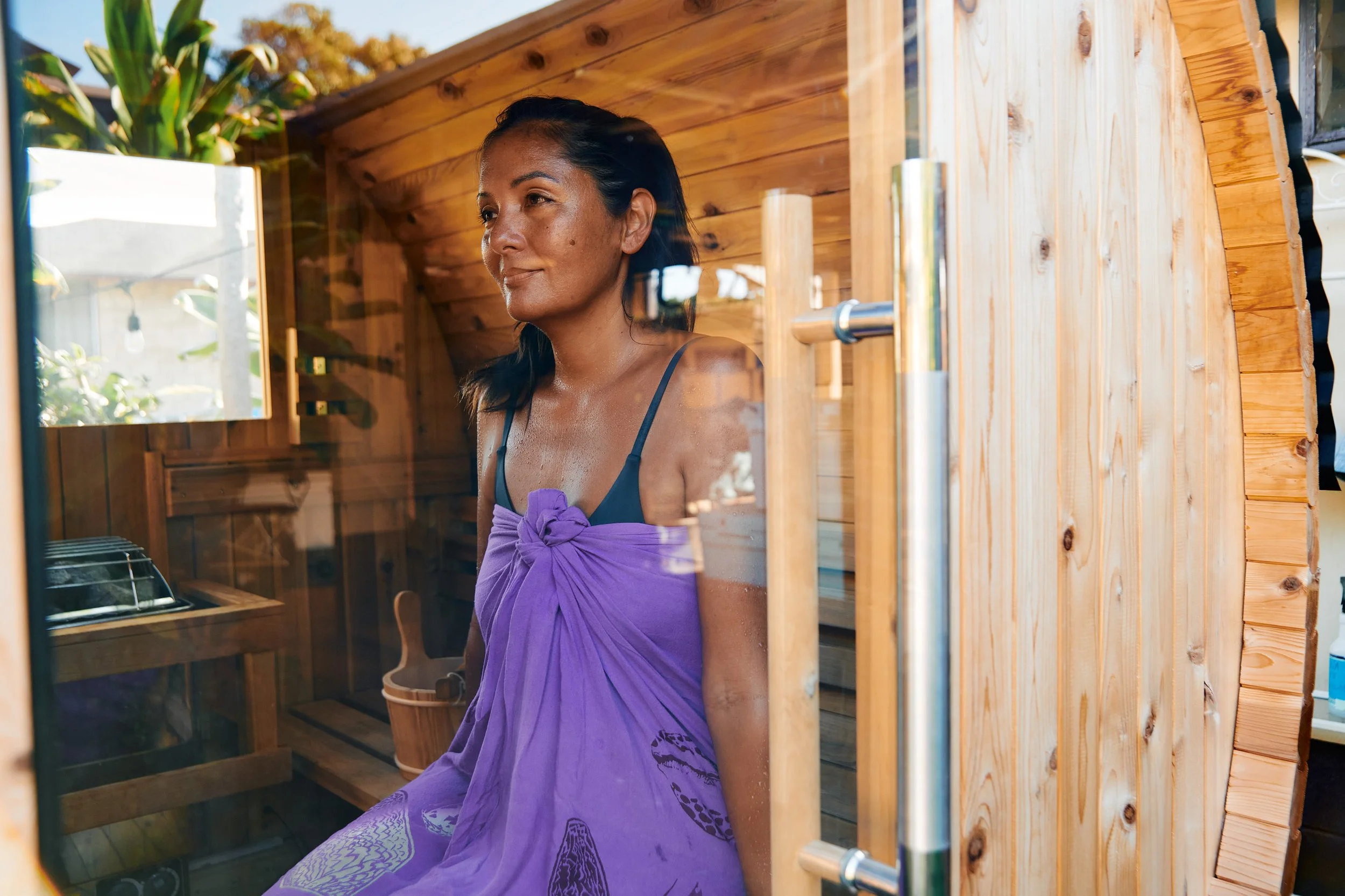 A woman with dark hair and a towel wrapped around her is relaxing inside a wooden sauna, looking out through glass with a content expression.