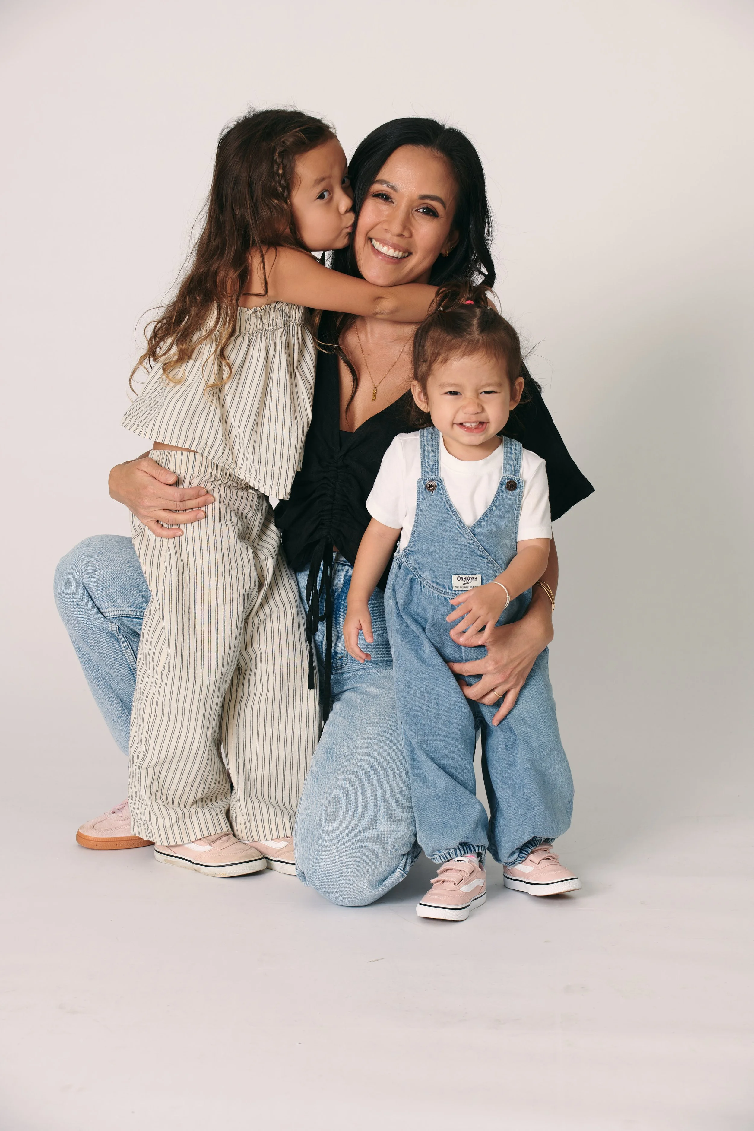 A woman smiling and kneeling with two young girls, one giving her a kiss on the cheek and the other standing in front with a smile, all dressed casually in light-colored clothes against a plain white background.