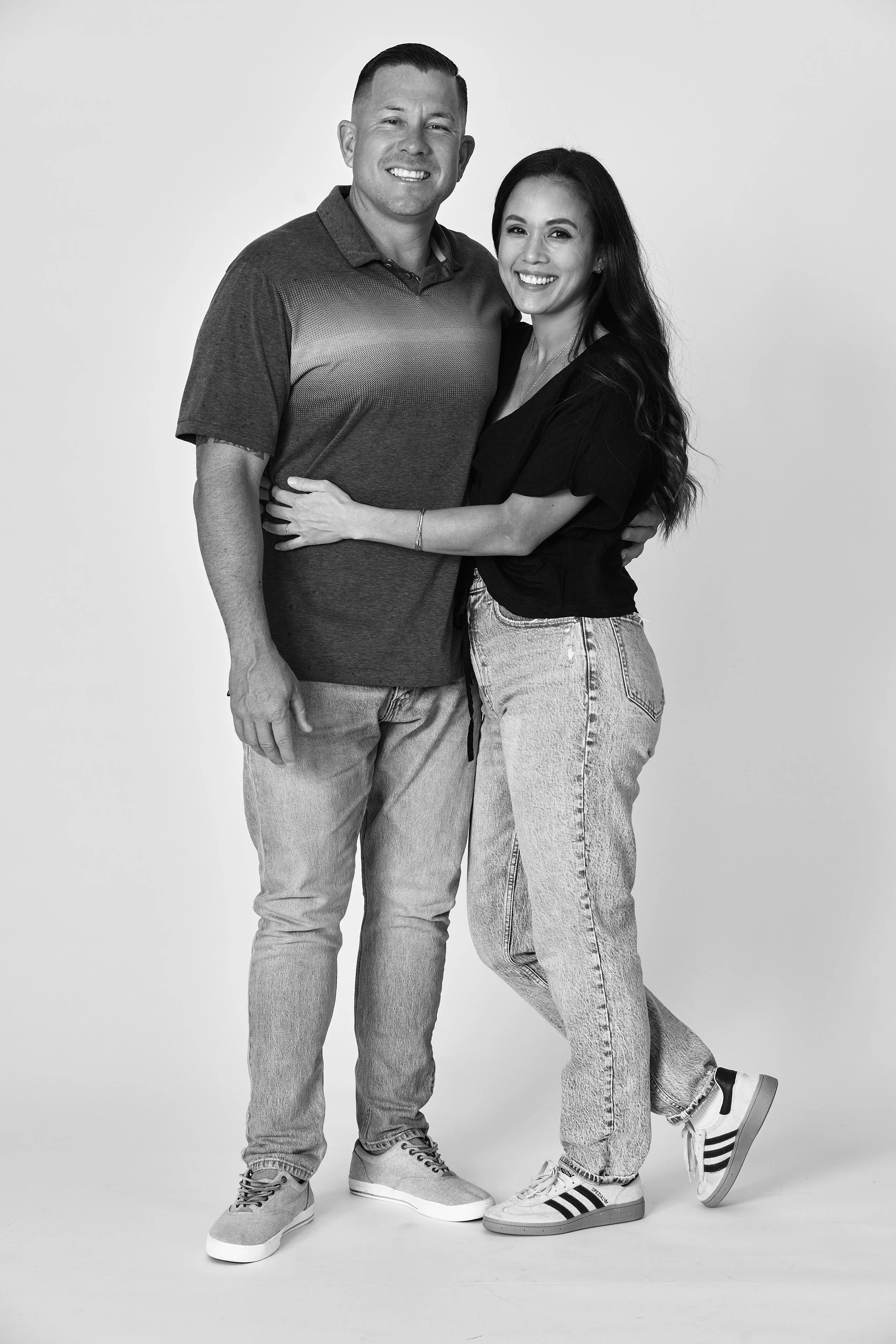 A black-and-white photo of a smiling couple standing close together and hugging. The man is wearing a short-sleeved polo shirt, jeans, and sneakers. The woman is wearing a black top, high-waisted jeans, and sneakers.