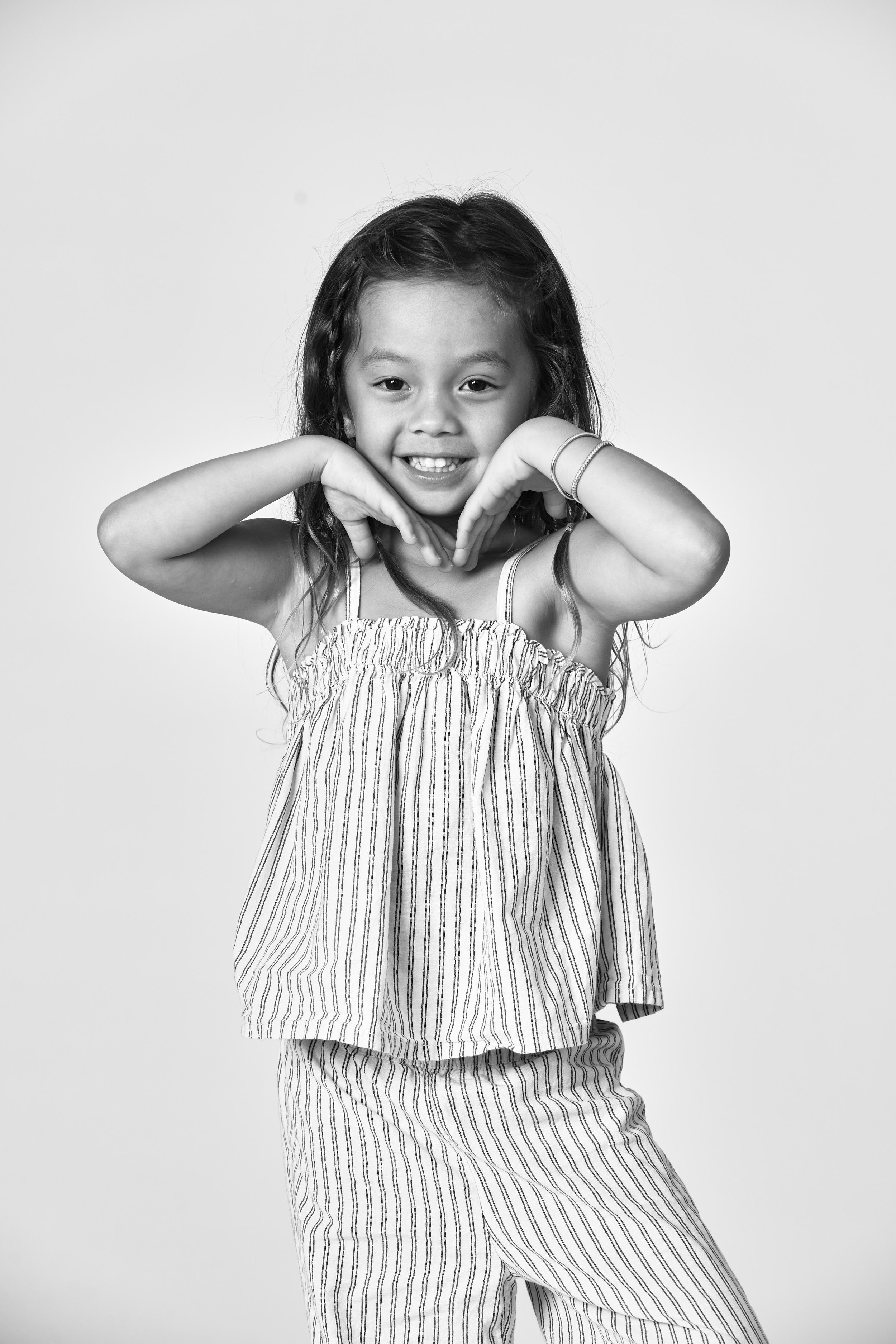 A young girl with long hair smiling and making a heart shape with her hands in front of her face, photographed in black and white against a plain background.