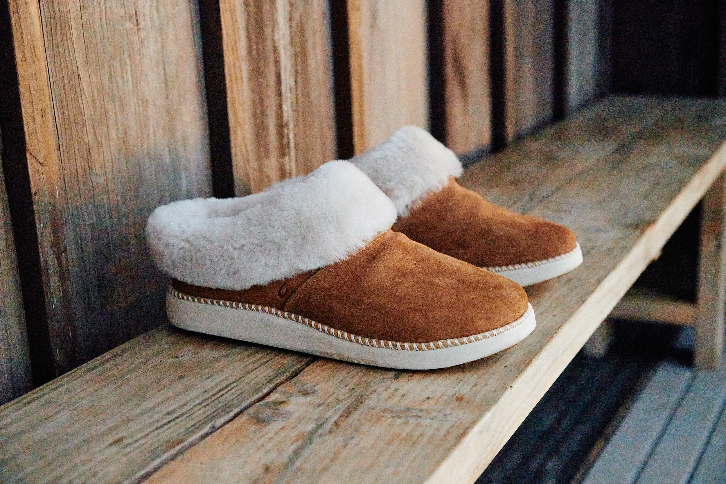 A pair of brown and white slippers with plush white lining on a wooden shelf against a wooden wall.