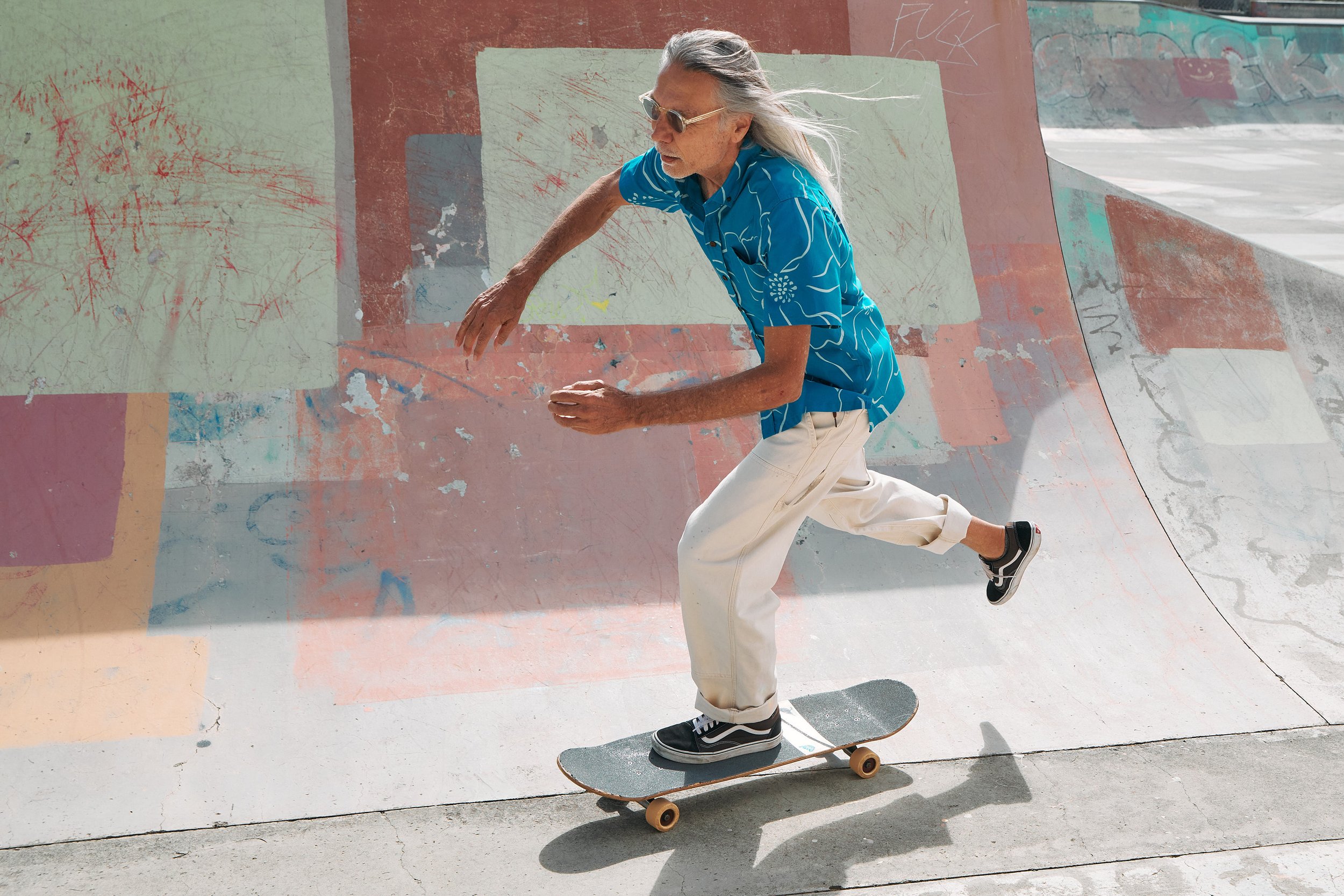 An elderly man with long gray hair, wearing sunglasses, a blue patterned shirt, and white pants, skateboarding in a skatepark with colorful graffiti and abstract murals.