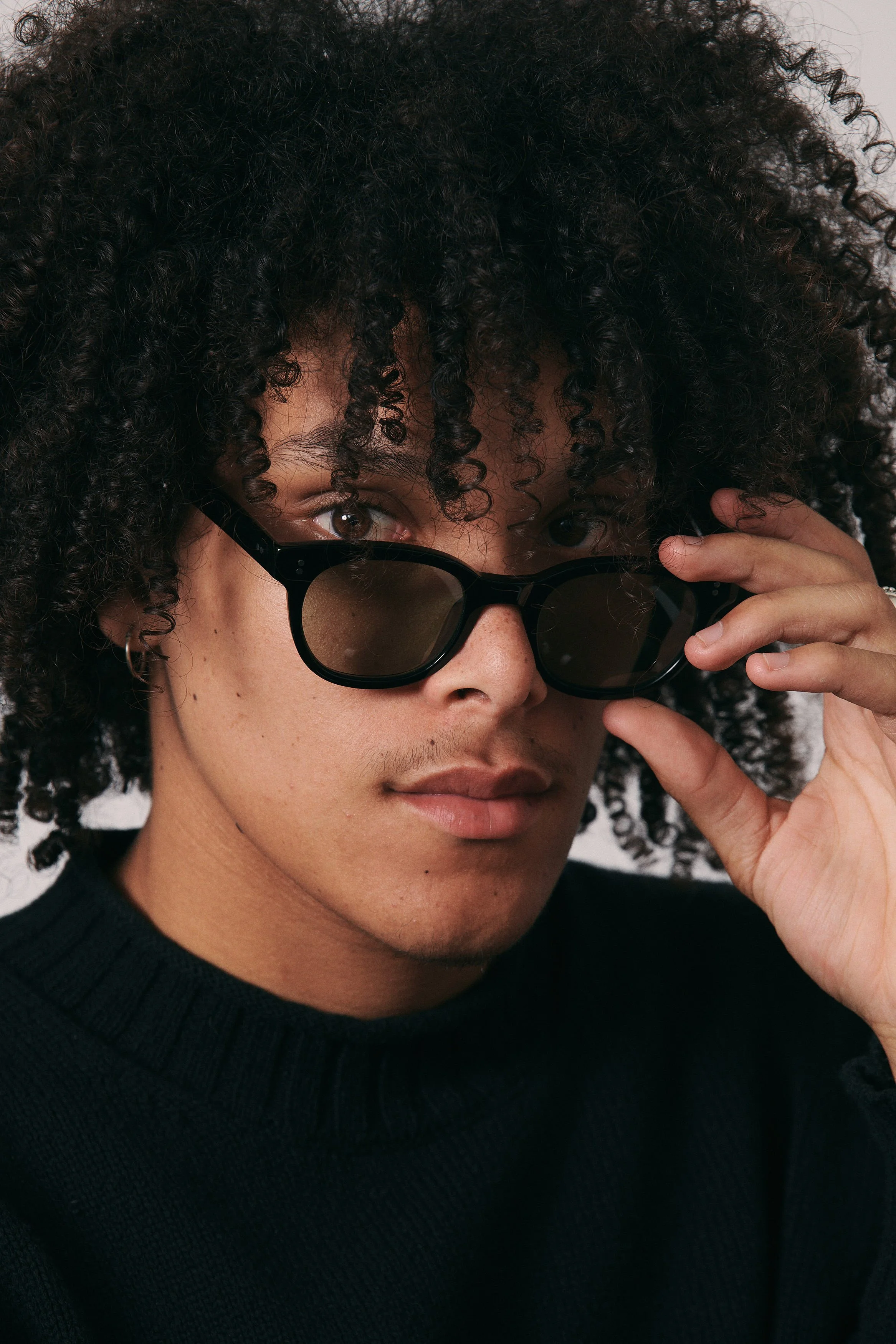 A person with curly hair wearing sunglasses and a black sweater, looking at the camera.