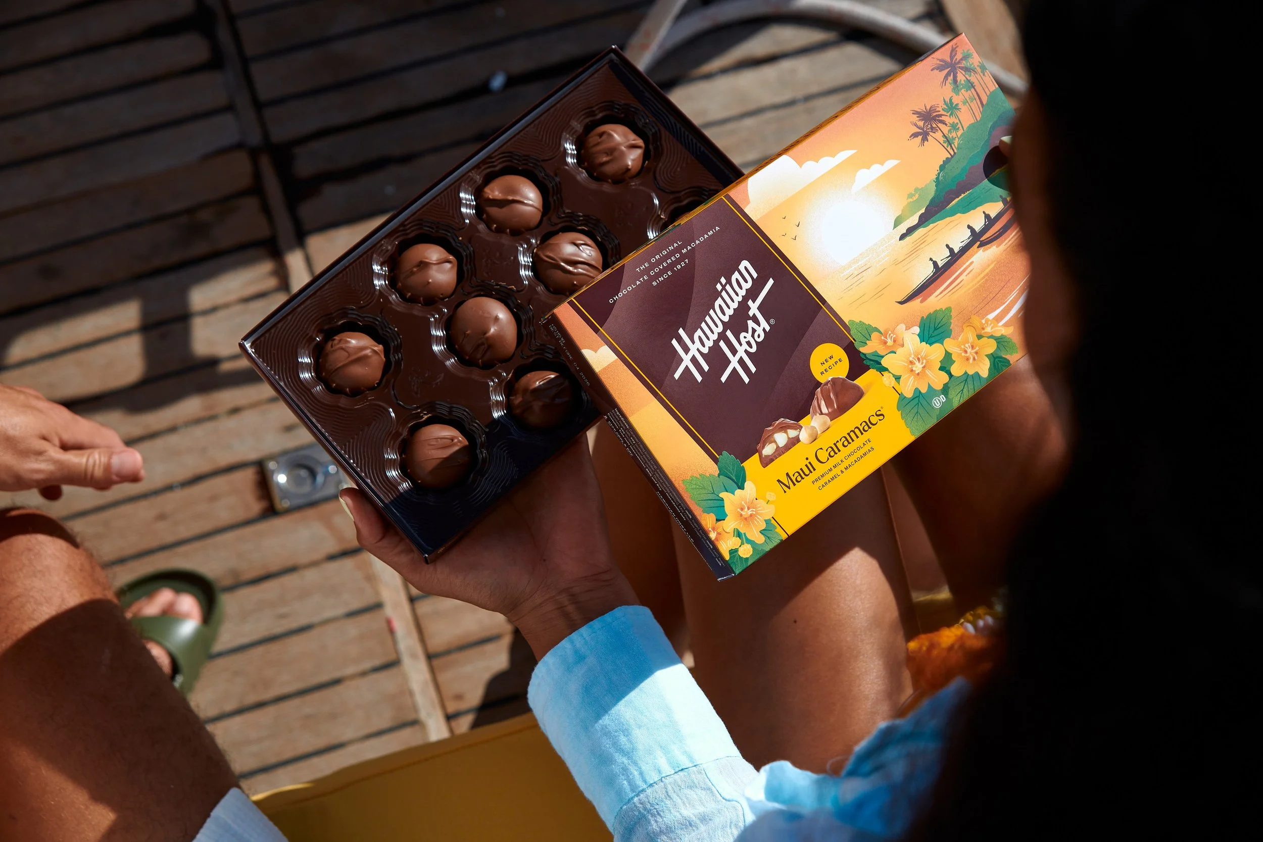 A person holding a box of Hawaiian Host Maui Caramacs chocolates with a partly open box revealing milk chocolates on a wooden deck.