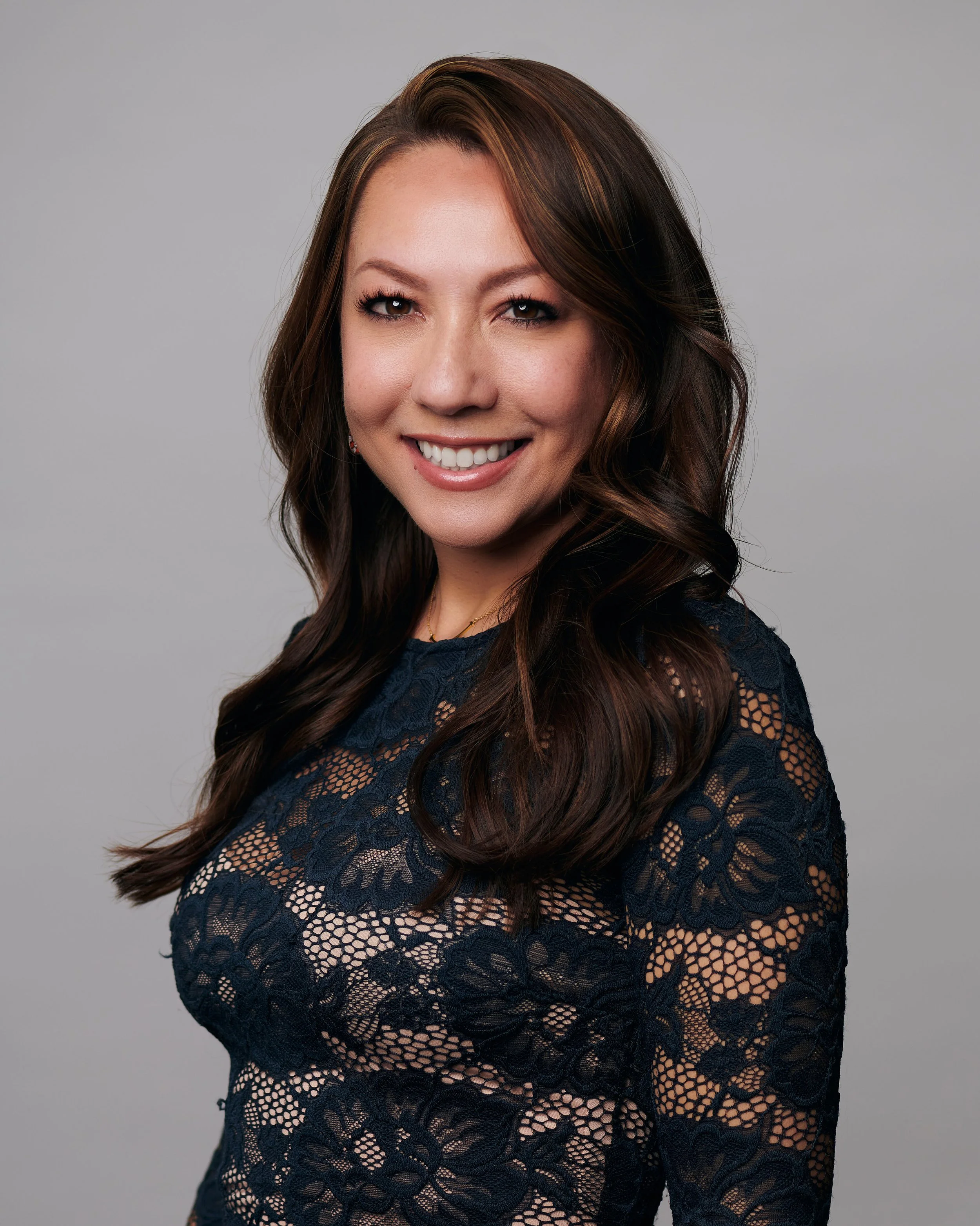 A woman with long wavy brown hair and fair skin smiling, wearing a navy lace top, against a neutral gray background.
