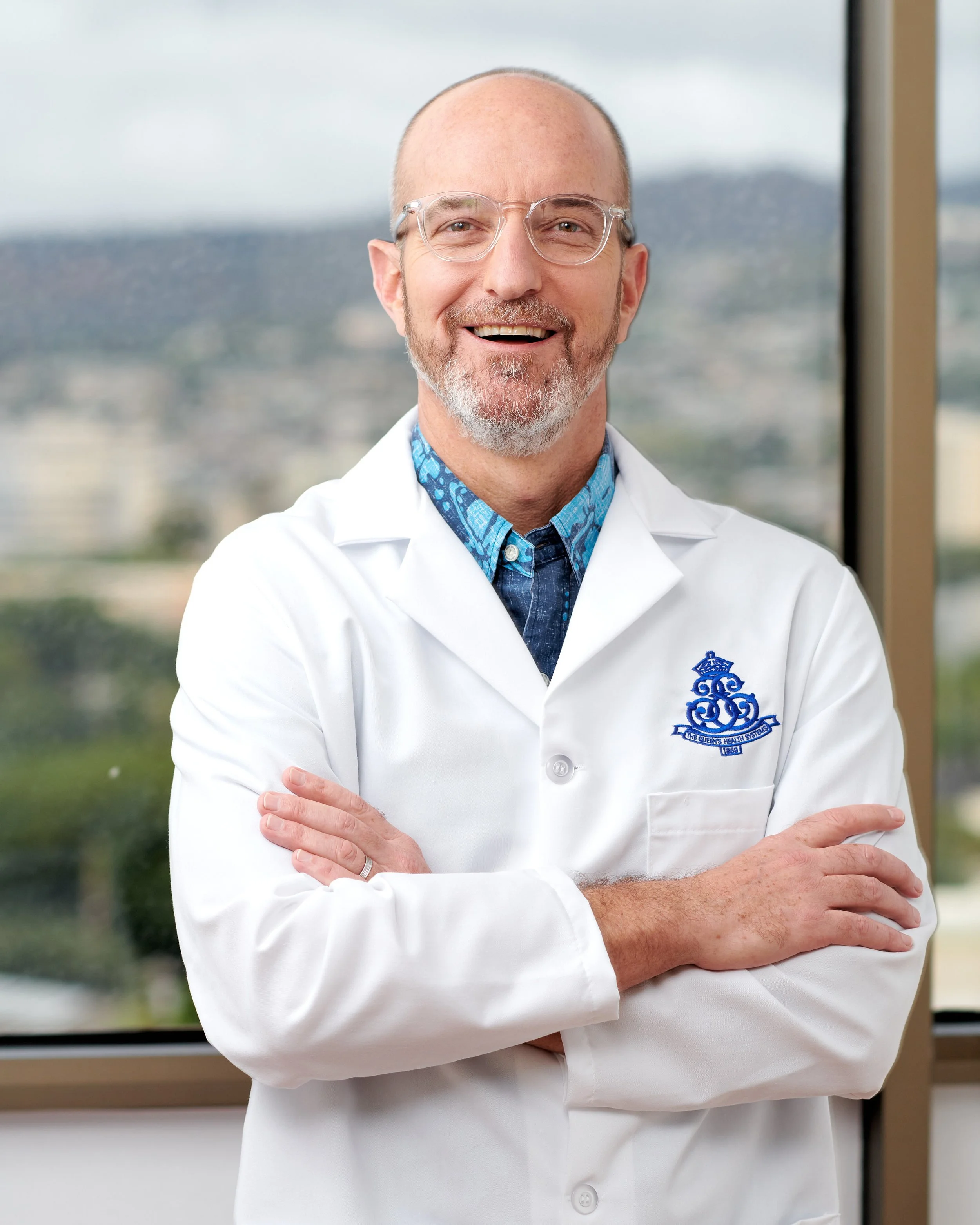 A smiling middle-aged man with glasses and a beard wearing a white lab coat standing in front of a large window with a cityscape view.