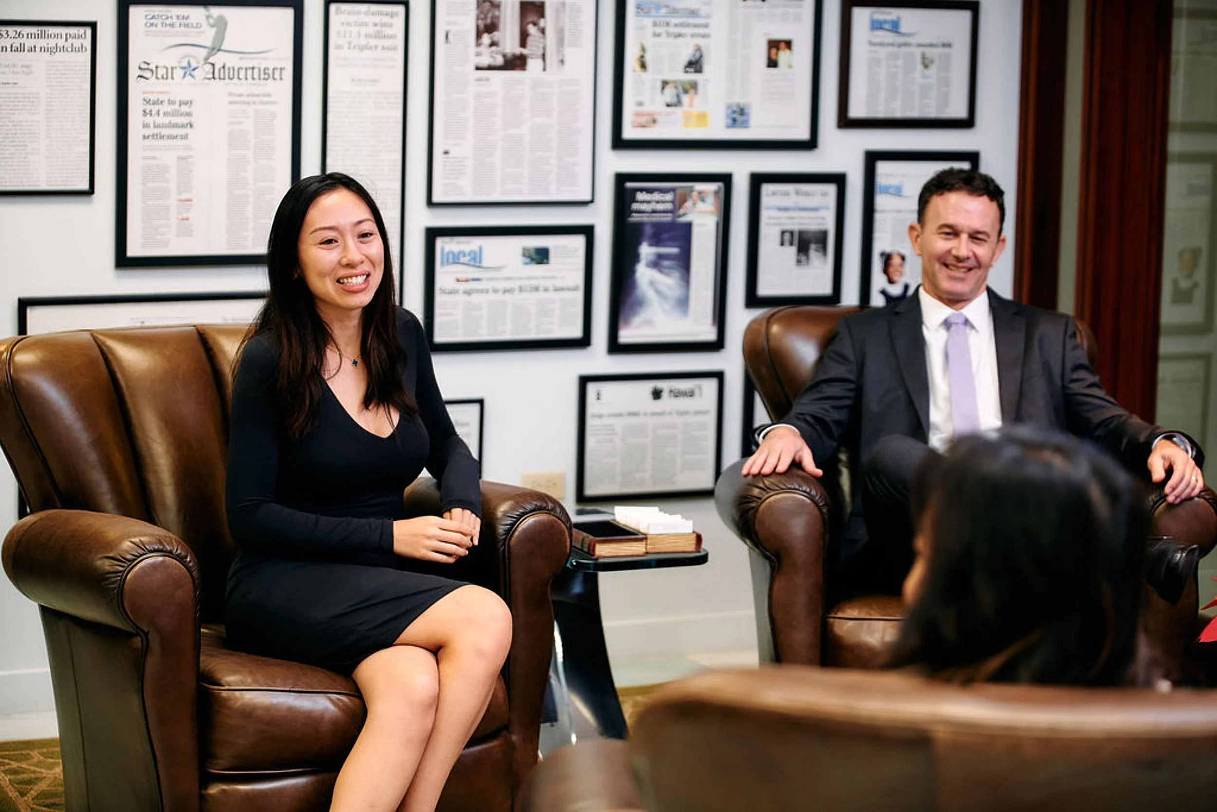 Two people, a woman and a man, sitting on leather armchairs in a formal setting. The woman has long black hair, is wearing a black dress, and is smiling. The man has short hair, is wearing a suit with a light-colored tie, and is also smiling. There a