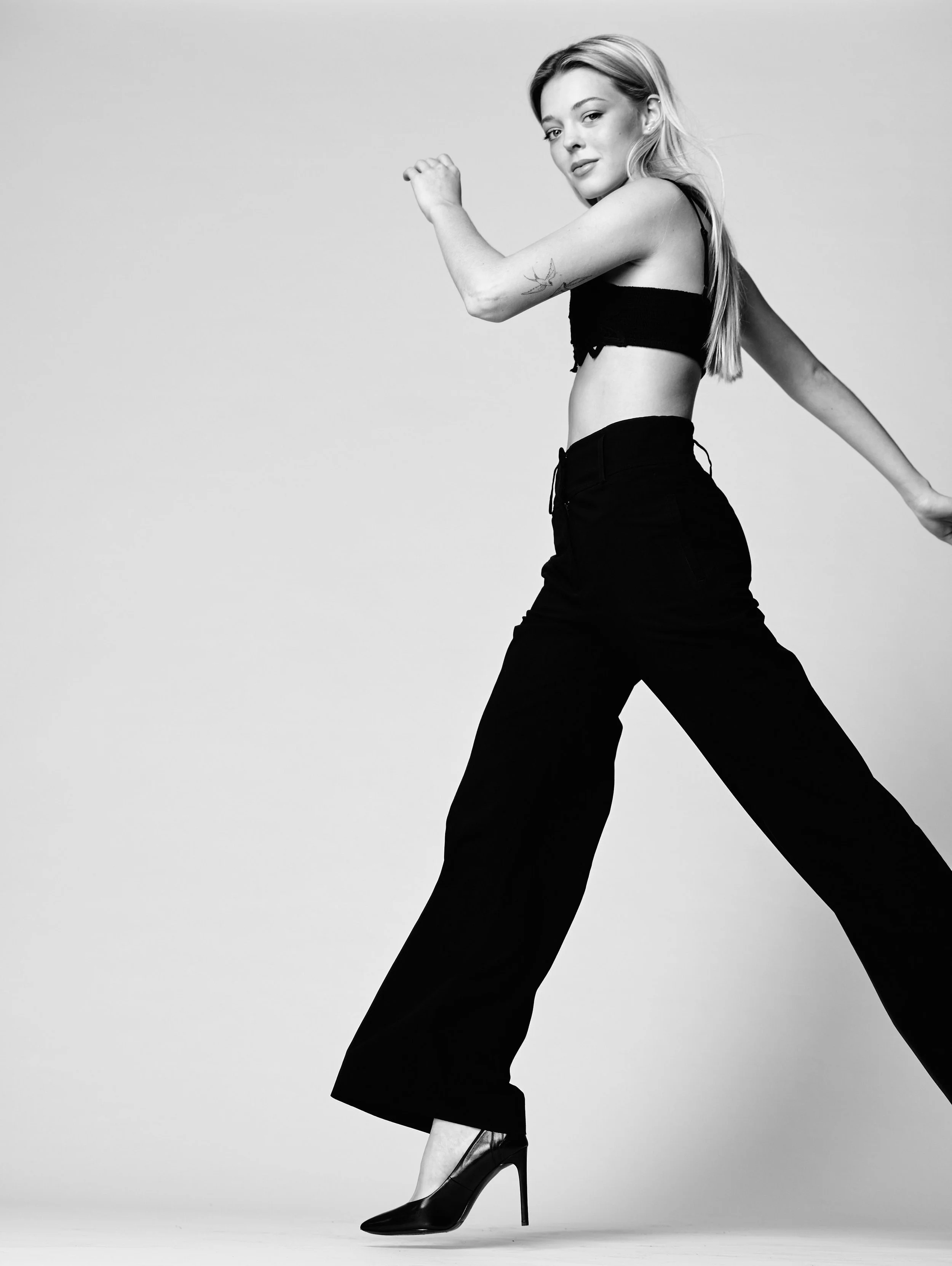 Black and white photo of a woman with long hair, wearing high heels, a crop top, and high-waisted pants, posing with one hand raised and the other arm extended behind her.