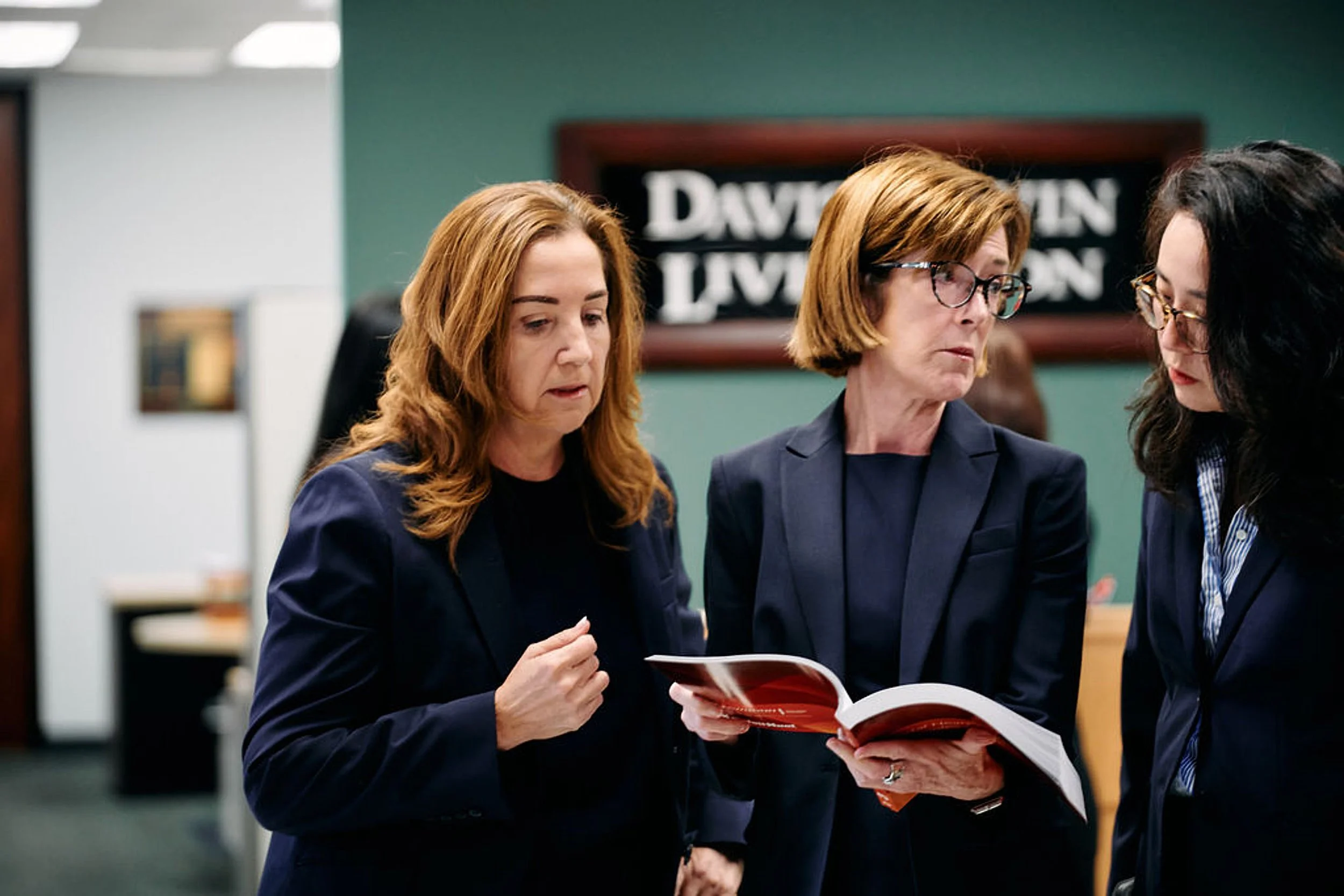 Three women in business attire in an office, examining a document or book together.