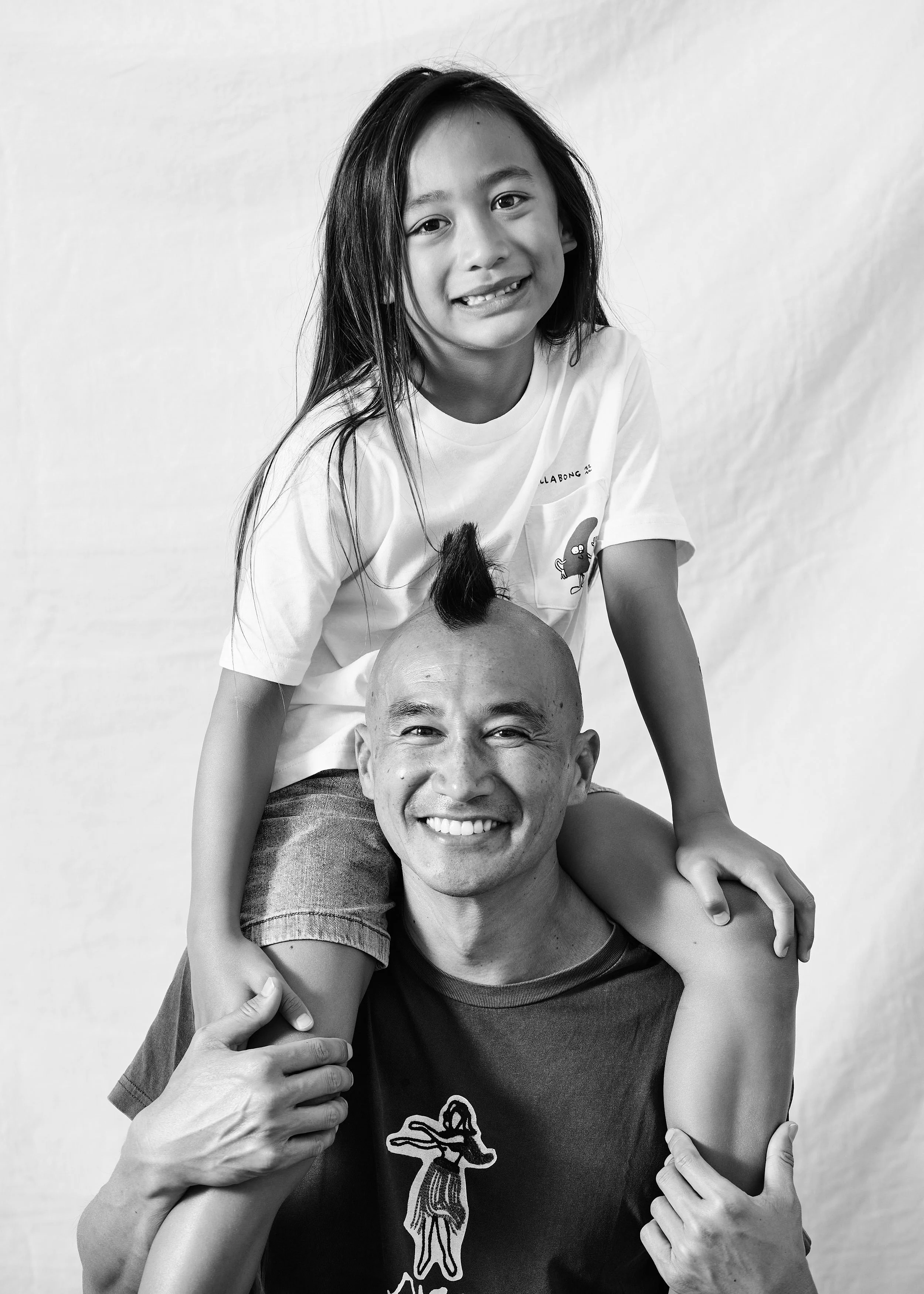 A man with a shaved head and a smile, holding a young girl with long dark hair on his shoulders. The girl is smiling and has a playful mohawk hairstyle. Both are wearing casual T-shirts and are posed against a plain background, captured in black and 