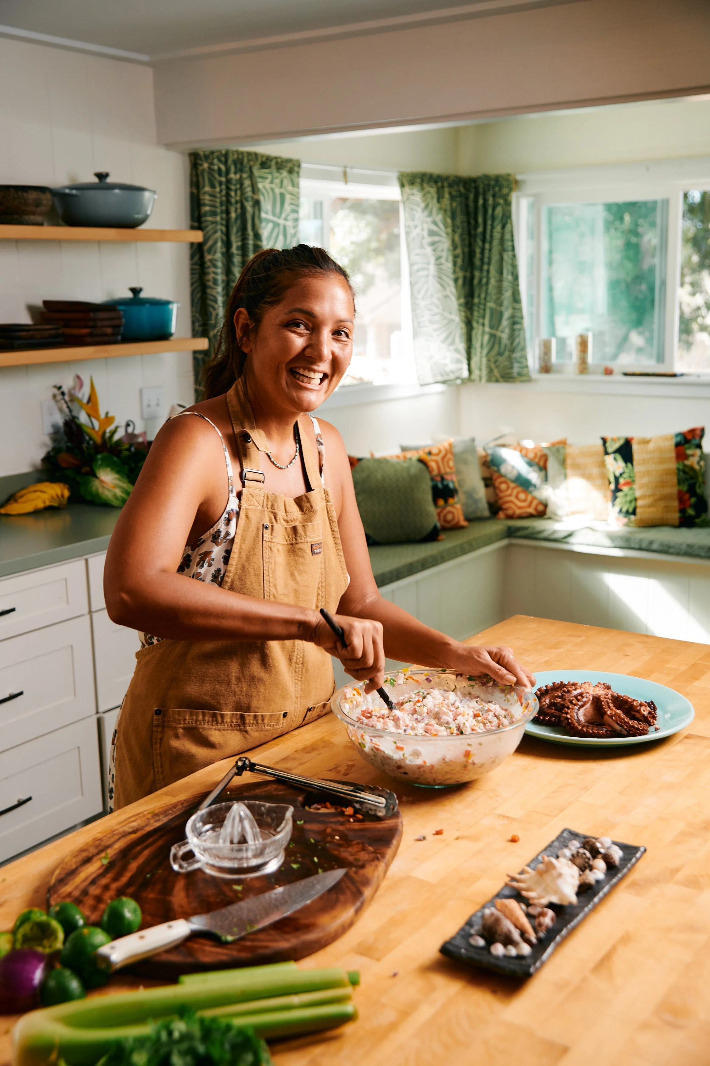 A woman in an apron is preparing food in a bright kitchen, smiling at the camera. The counter has vegetables, a knife, a bowl, and a plate of cooked octopus.