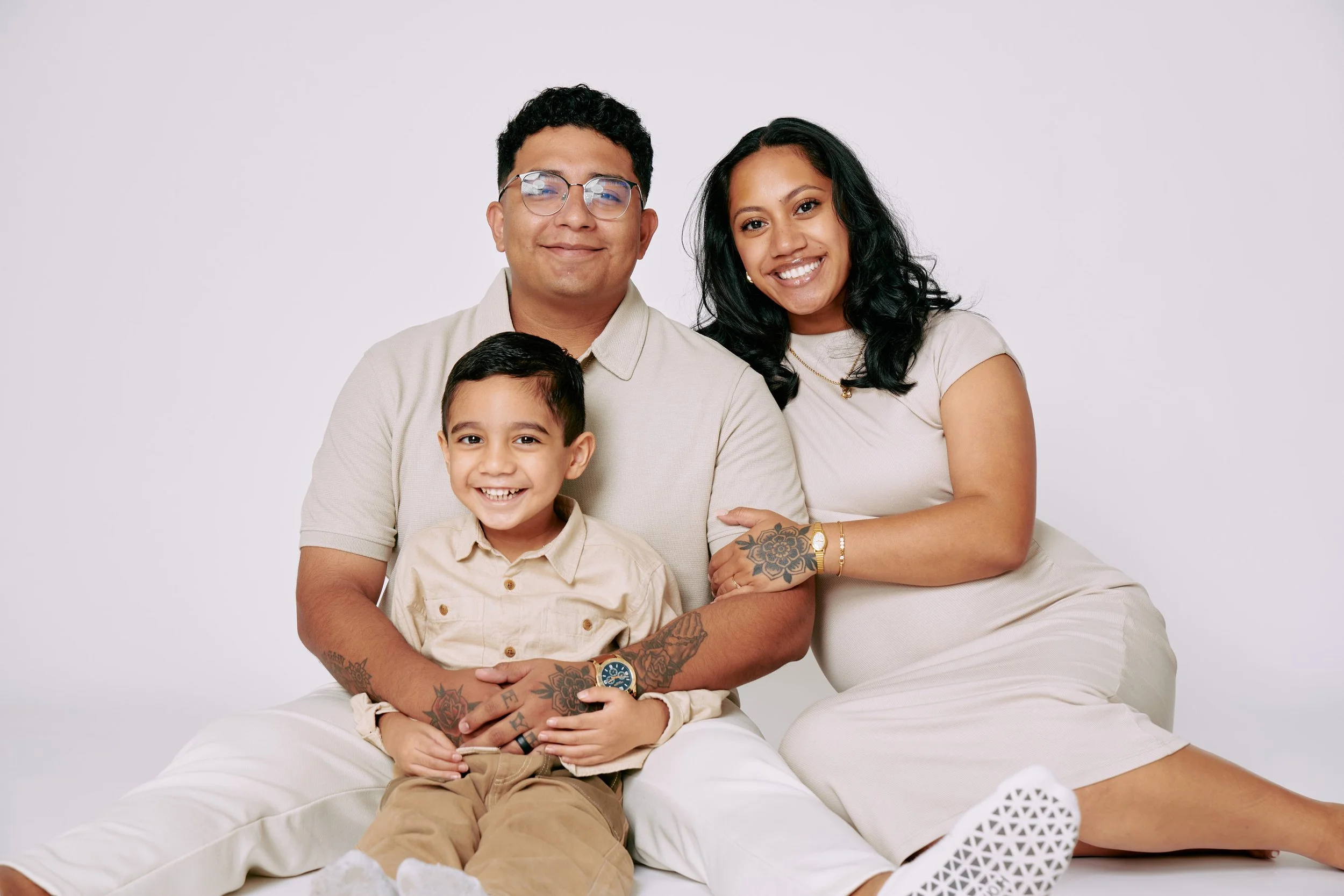A happy family of three sitting together against a plain white background, consisting of a smiling woman with dark curly hair, a man wearing glasses, and a young boy with short dark hair.