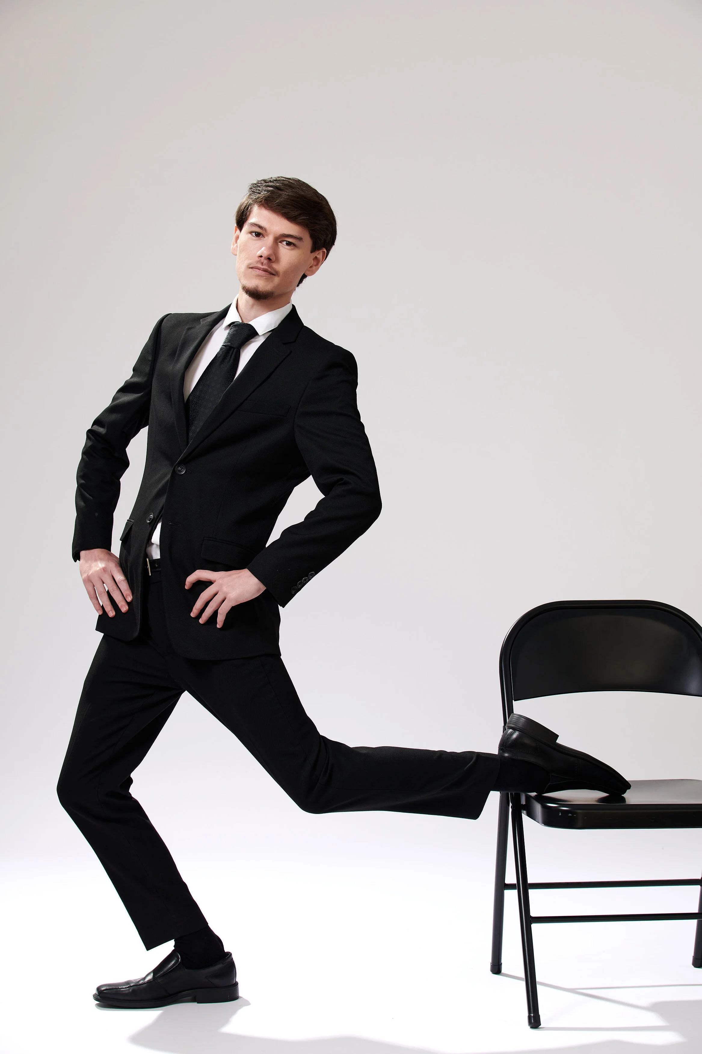 A man in a black suit with a white shirt and black tie, stretching his leg on a black chair, standing against a plain background.