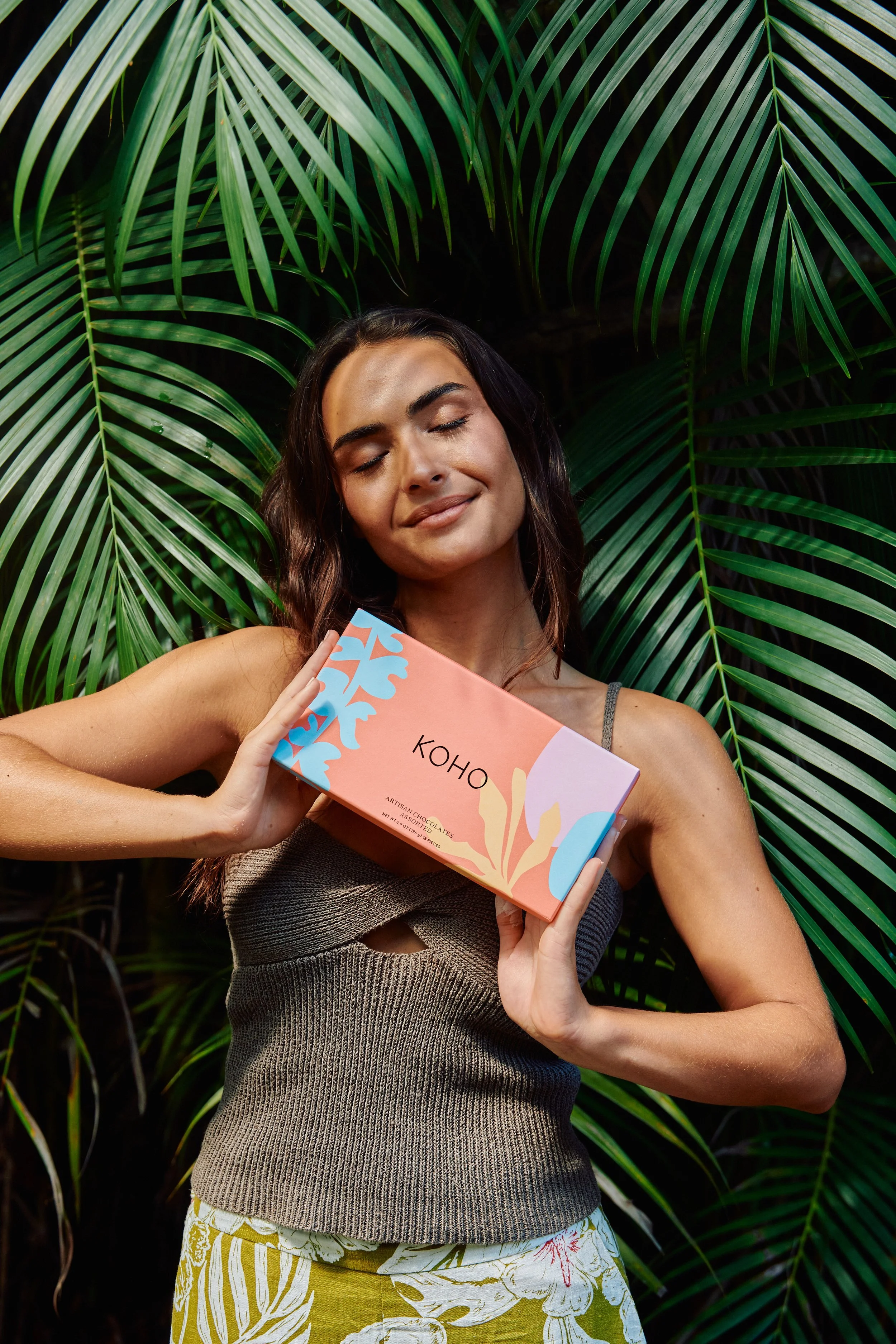 A woman with closed eyes holds a pink box labeled 'KOHO' close to her face while standing amidst large green palm leaves.