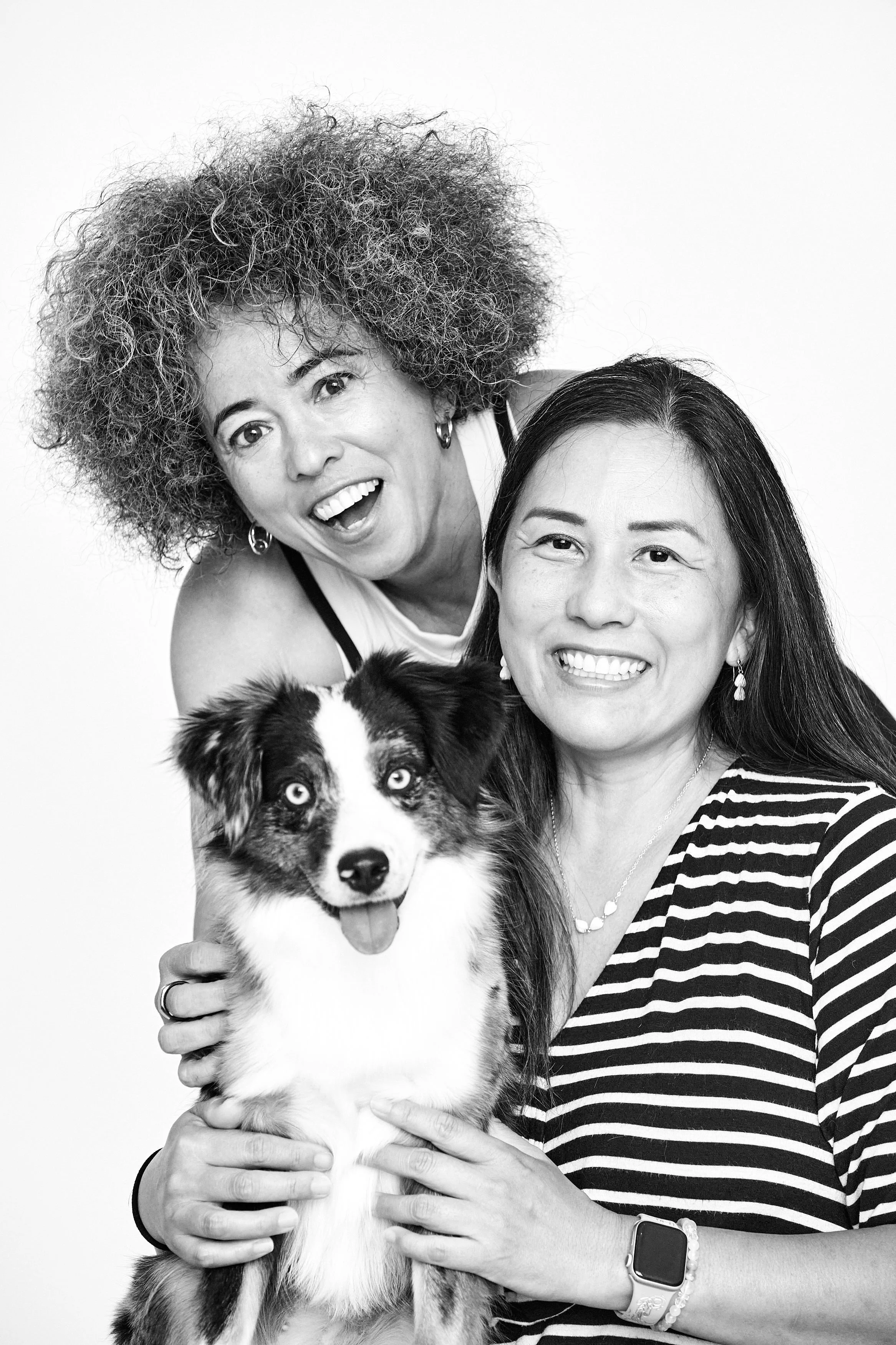 Black and white photo of two women smiling and holding a dog with a white and black coat and striking blue eyes.