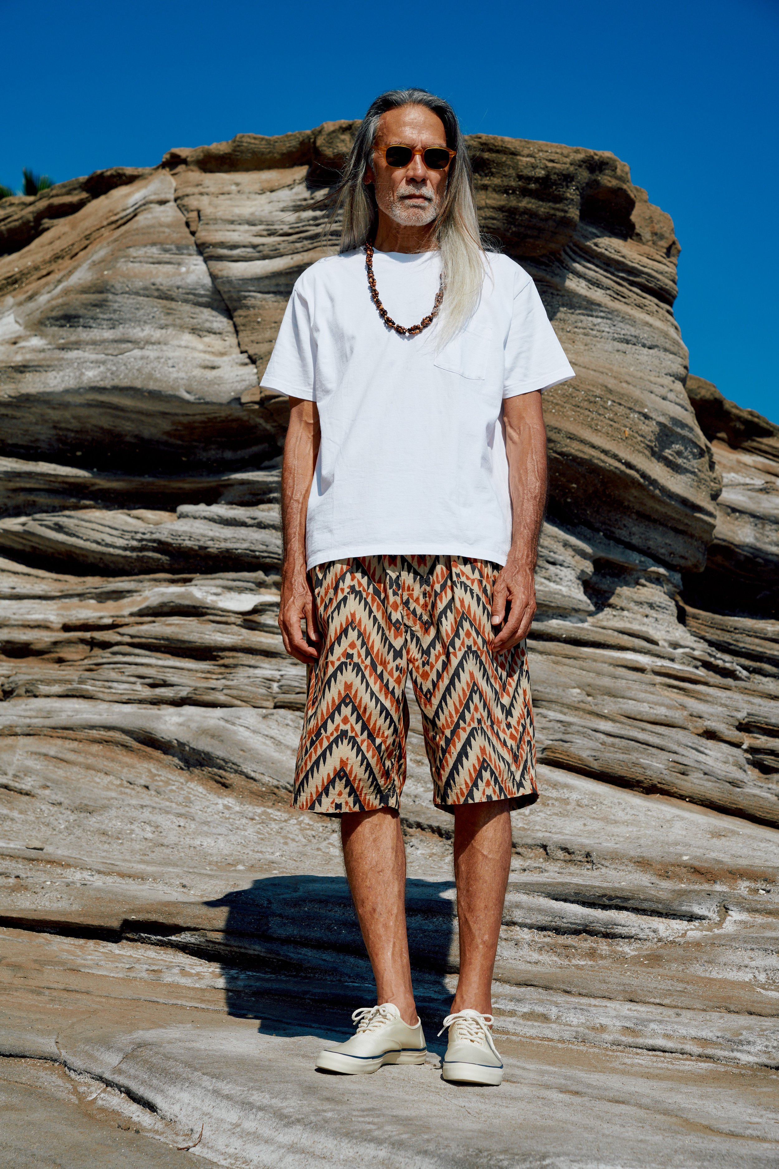 An older man with long gray hair, sunglasses, and a white T-shirt standing outdoors on rocky terrain with a clear blue sky in the background.