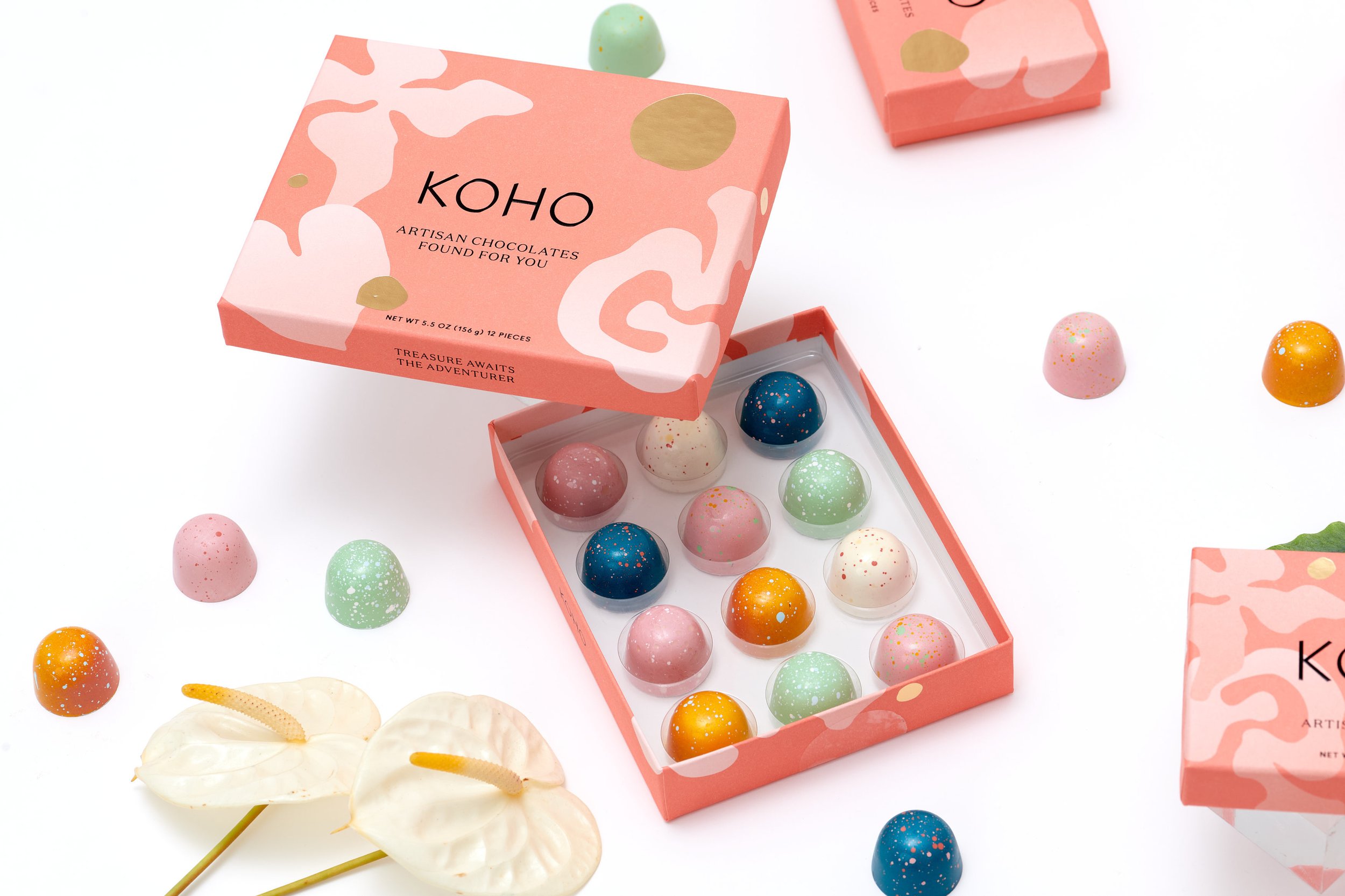 Open pink box of Koho artisan chocolates with various colorful, speckled chocolates inside, and some outside the box on a white surface. Two white calla lilies are also seen in the bottom left corner.