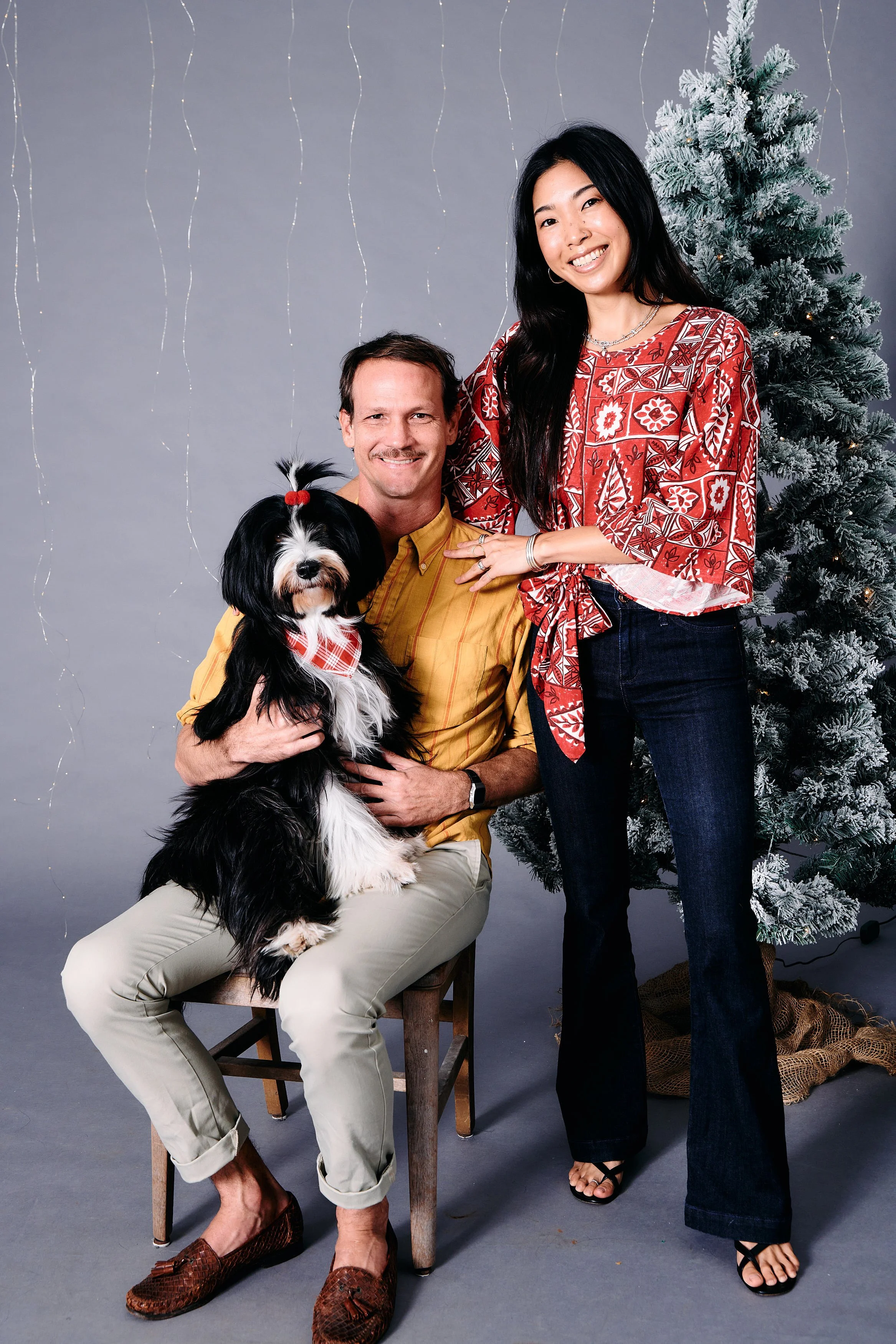 A man, woman, and dog pose in a Christmas-themed photo. The man is seated on a wooden chair, holding a black and white dog with a red and white checkered bandana and a top knot. The woman stands beside him with her hand resting on his shoulder. There