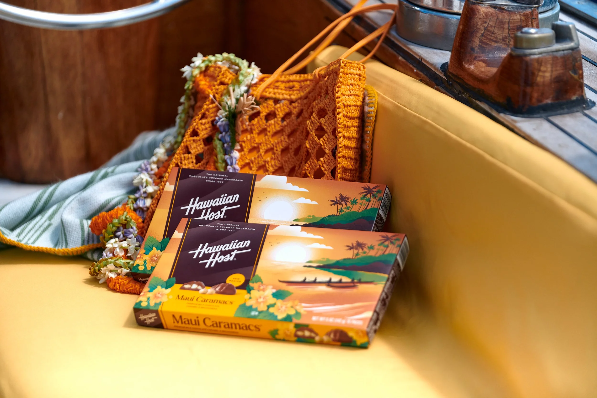 Two boxes of Hawaiian Host Maui Caramacs chocolates on a yellow table with leis, a crocheted bag, and wooden containers in the background.