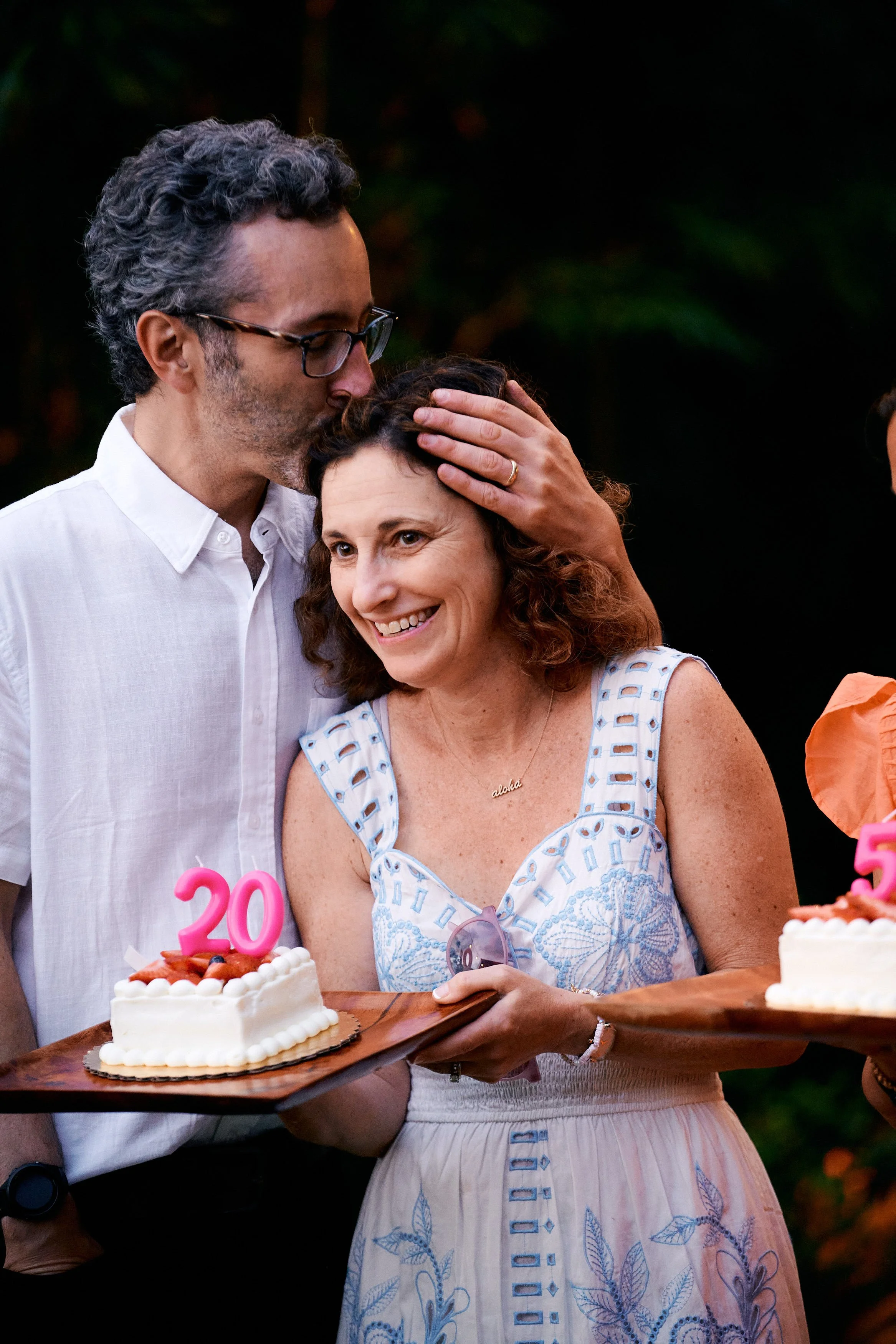 A man kisses a woman on the forehead during an outdoor celebration, holding a tray with a cake featuring a pink '20' candle.