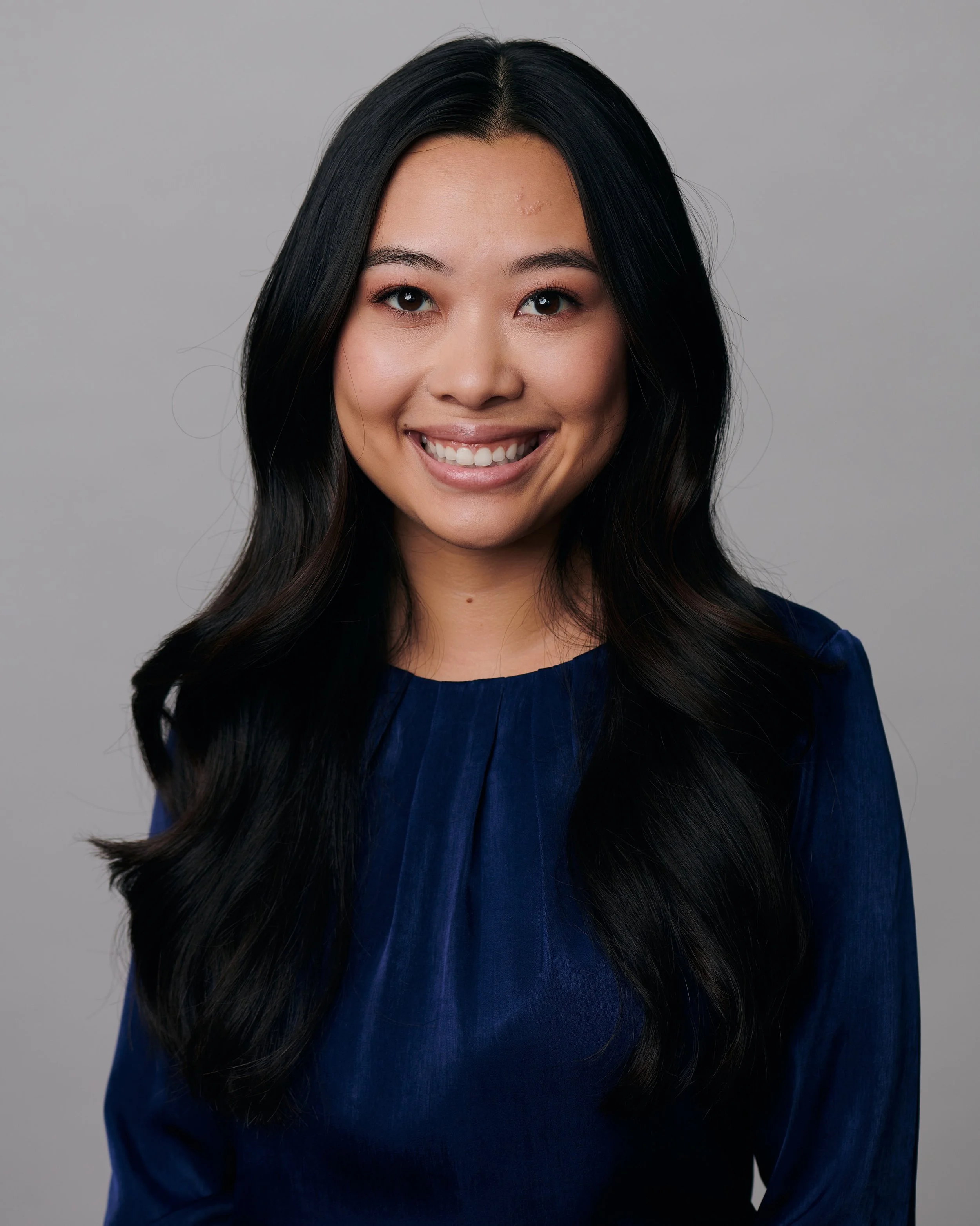 A young woman with long, dark hair smiling at the camera, wearing a dark blue top, against a plain gray background.