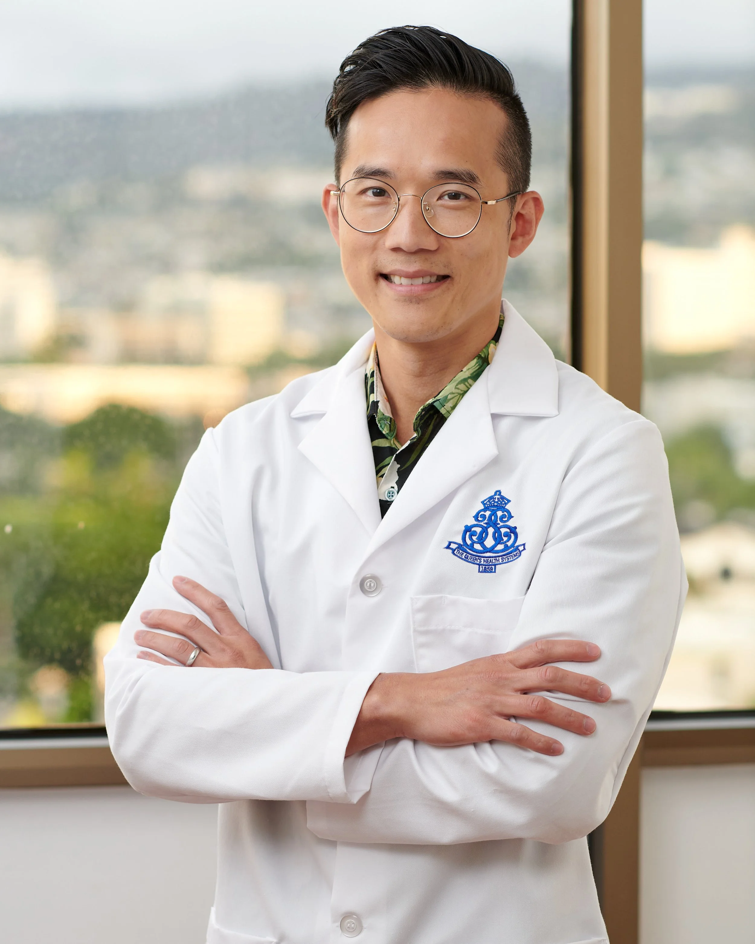 Young male doctor in white coat with crossed arms, wearing glasses, standing in front of a window with cityscape background.