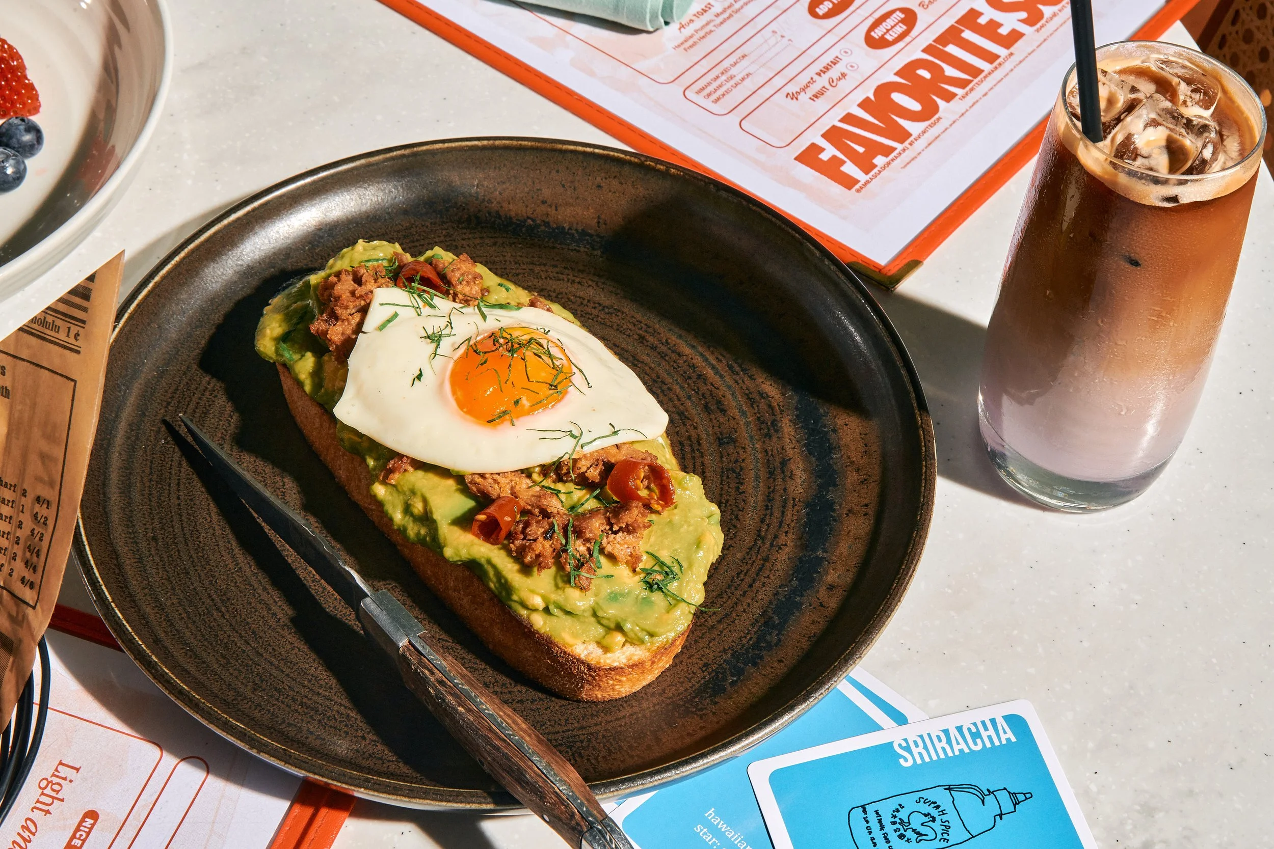A breakfast plate with avocado toast topped with ground meat, a fried egg, chopped tomatoes, and herbs, placed on a black plate. Next to it is a tall glass of iced coffee with a straw. The table has menus and a blue Sriracha sauce packet nearby.