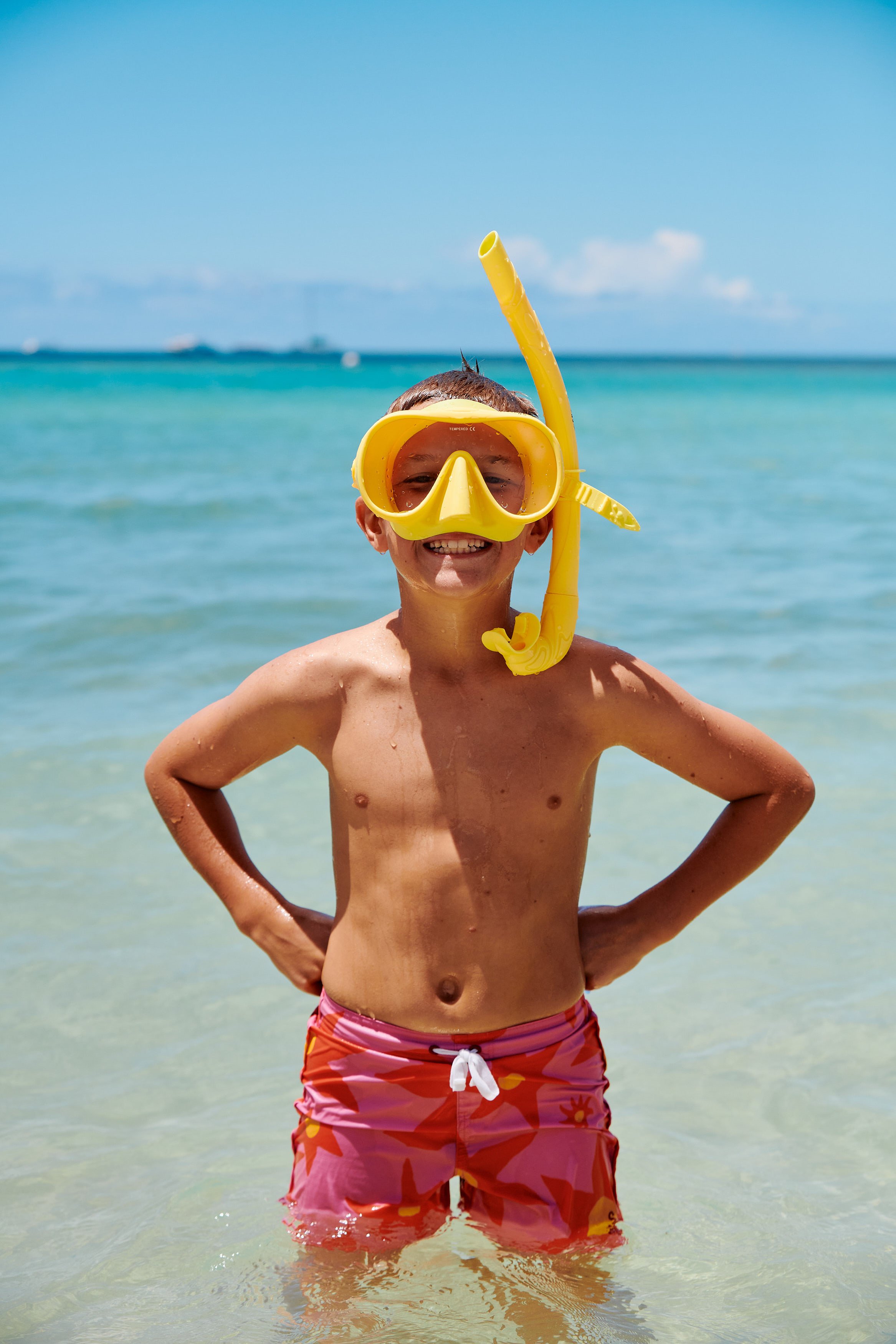 A smiling boy with a yellow snorkel and mask stands in shallow ocean water on a sunny day, wearing pink swim trunks with orange and yellow patterns.
