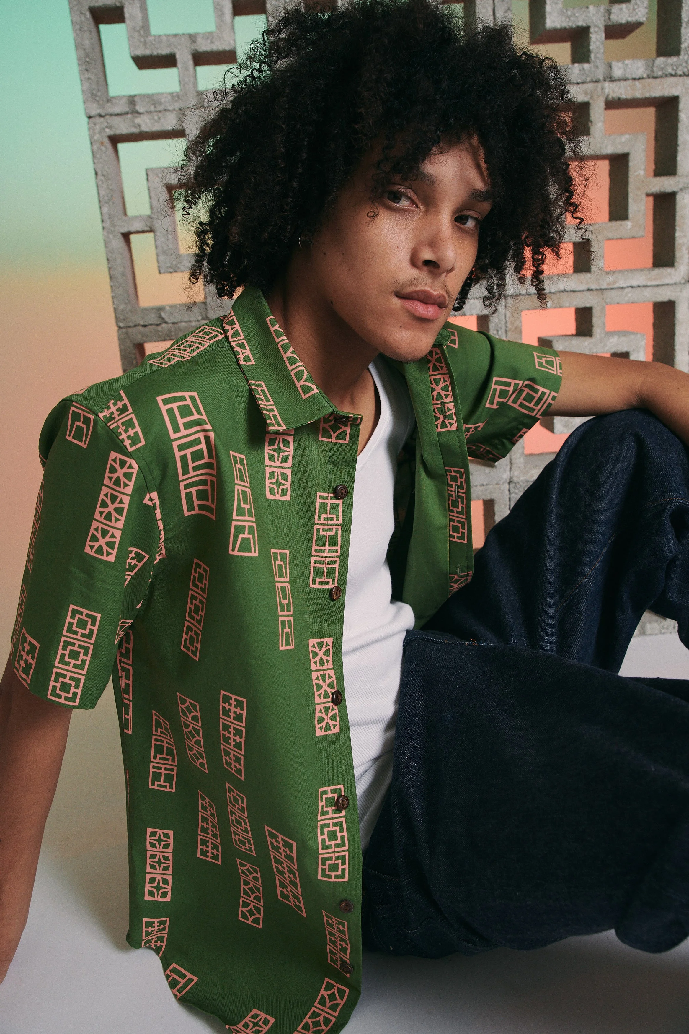 A young man with curly black hair wearing a green short-sleeved button-up shirt with pink geometric pattern, white undershirt, and dark jeans, sitting against a decorative concrete wall with geometric cutouts.