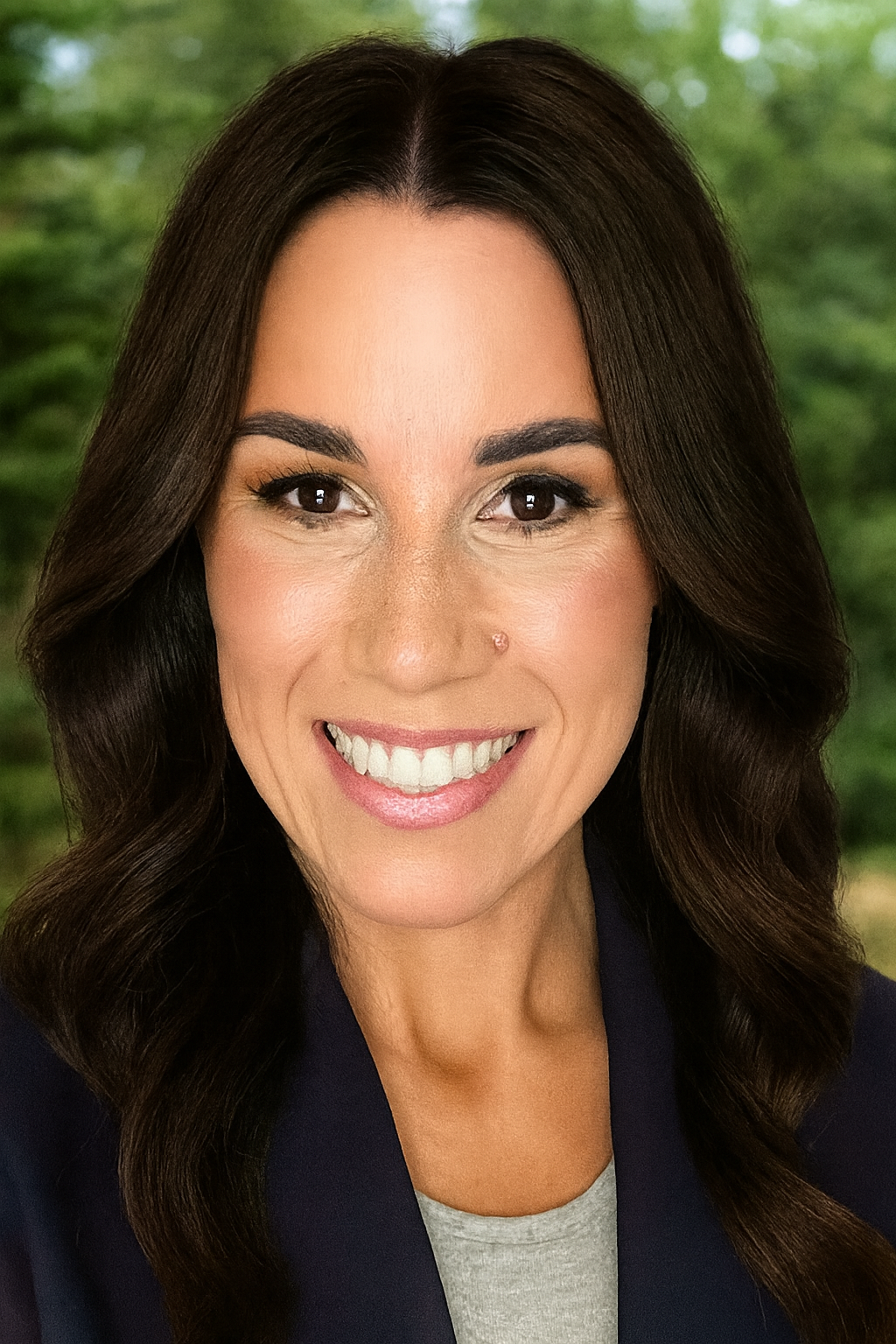 Close-up portrait of a woman with long dark wavy hair, brown eyes, and a bright smile, wearing a dark blazer and a light-colored top, with a blurred green outdoor background.