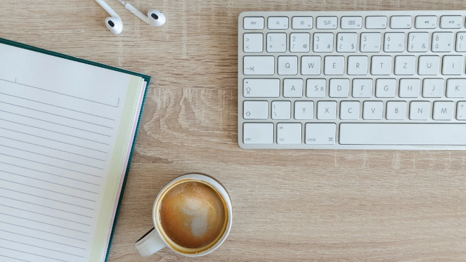 A tidy wooden desk seen from above, with an open lined notebook, a white keyboard, a cup of coffee, and a pair of earbuds.