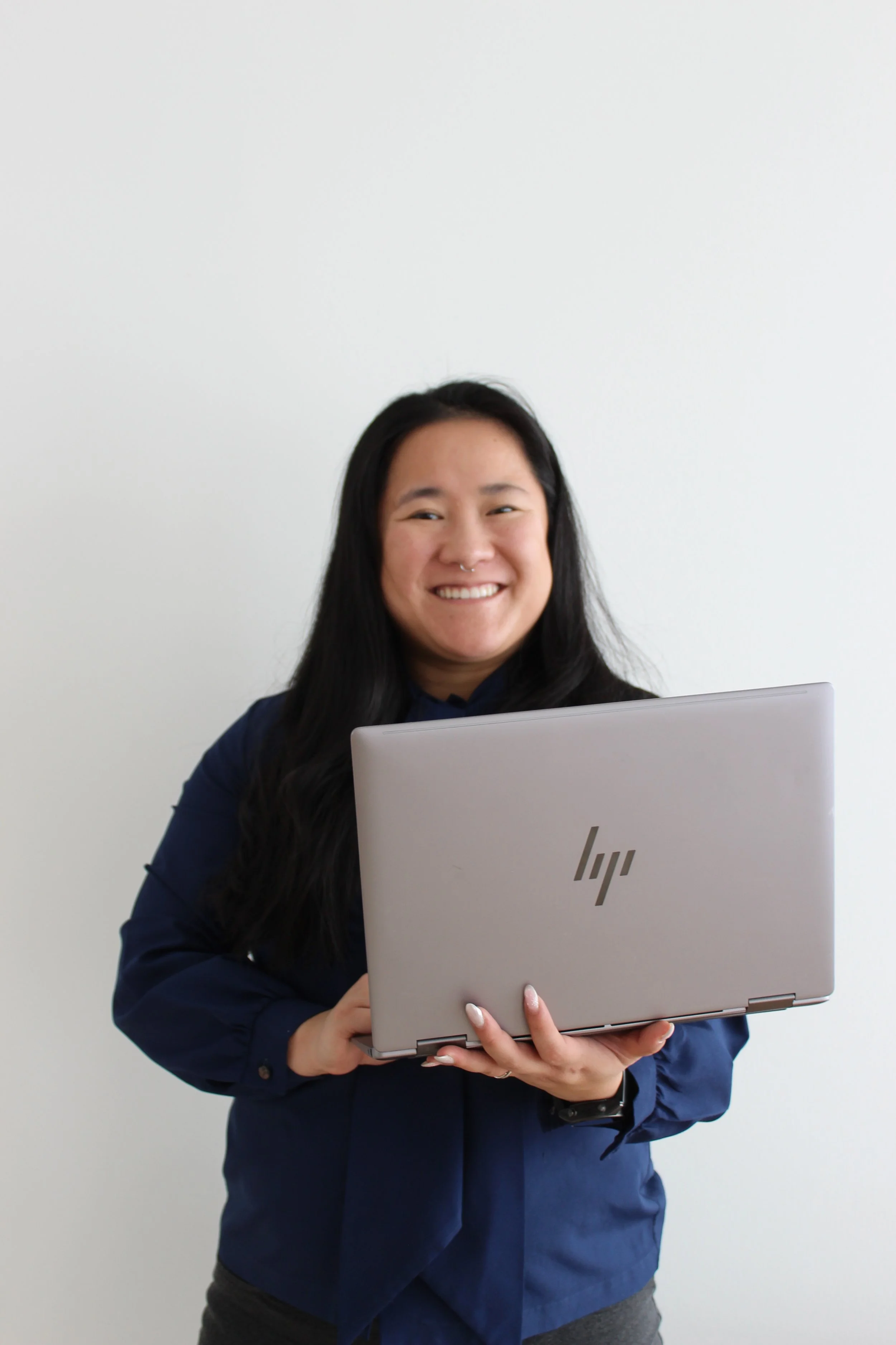 A woman with long black hair, wearing a dark blue blouse, is smiling while holding a silver HP laptop in front of a plain white wall.