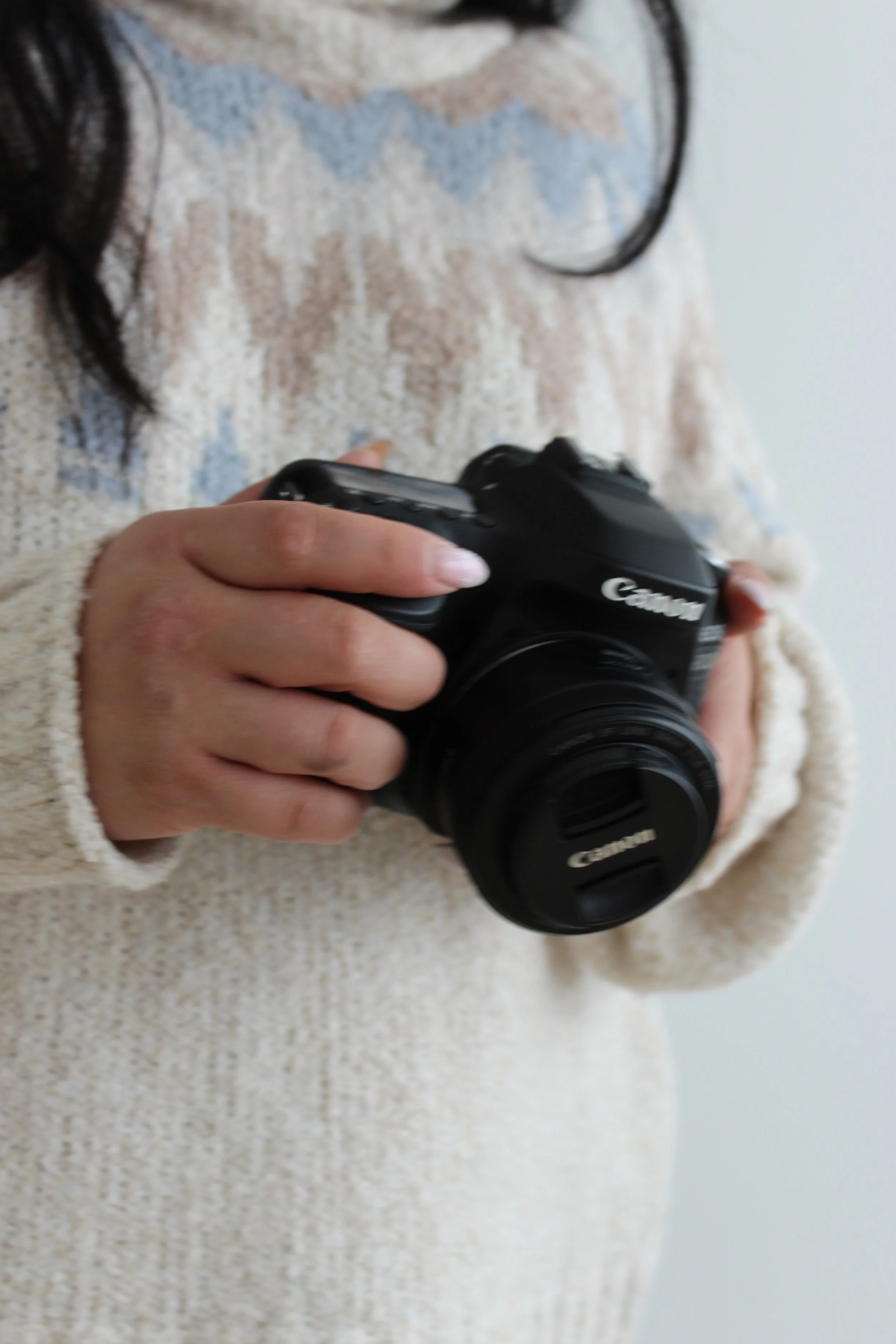 A person wearing a beige and multicolored sweater holding a black Canon camera.