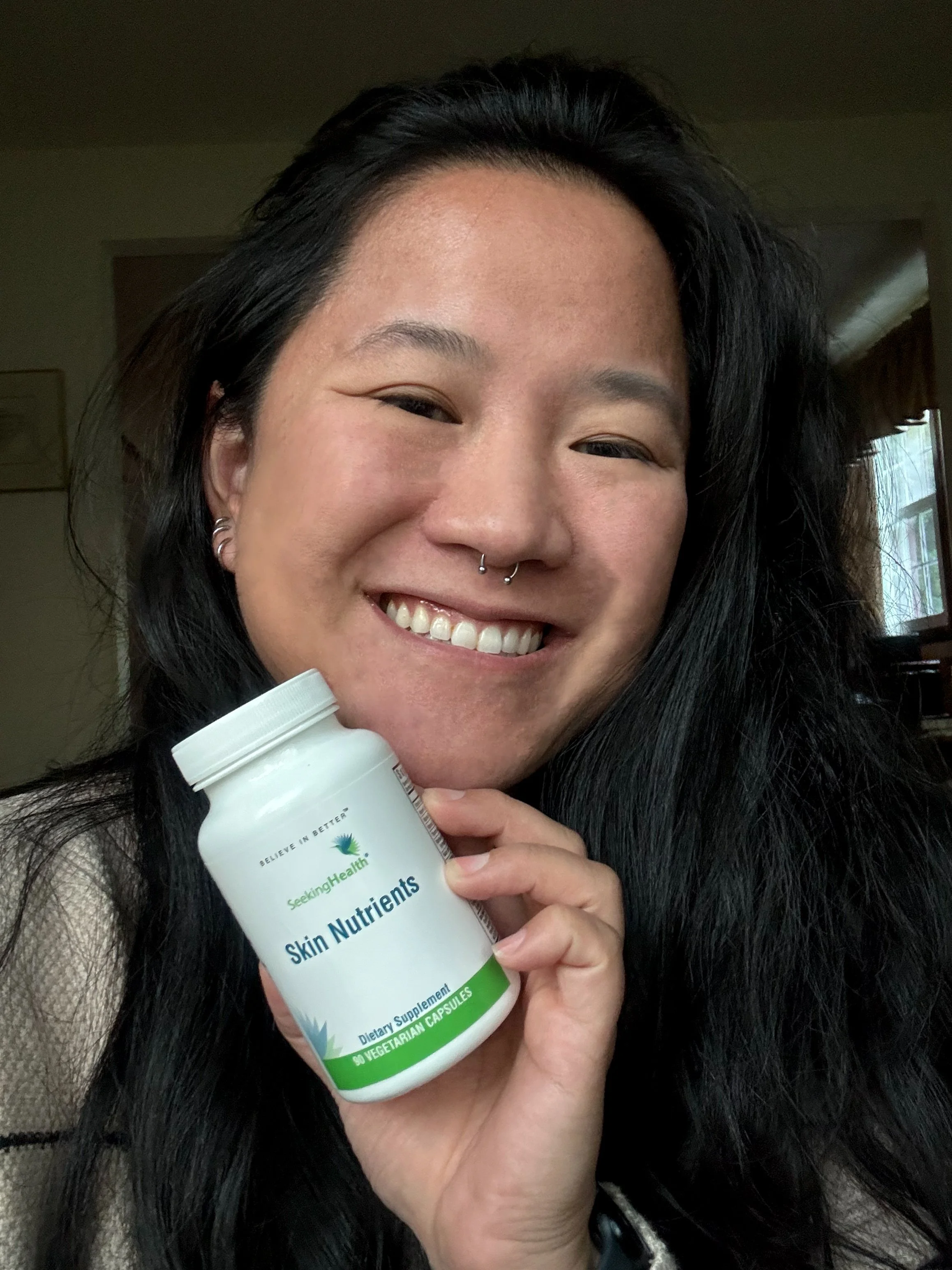 A smiling woman with long black hair holding a bottle of Skin Nutrients dietary supplements labeled Seeking Health.