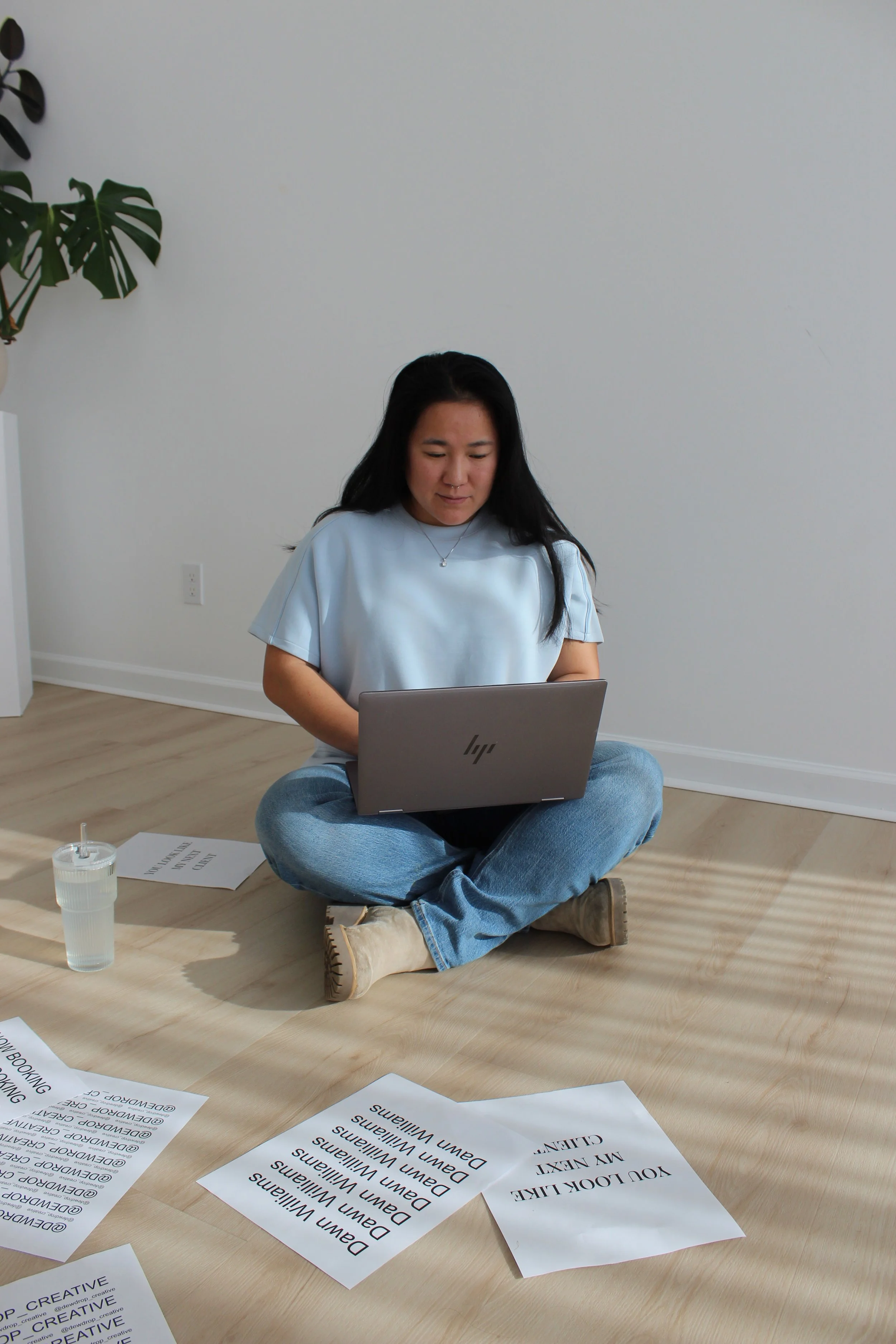 A woman sitting cross-legged on the floor using a laptop, with printed sheets of paper and a glass of water around her in a minimalist room with white walls and wooden flooring.