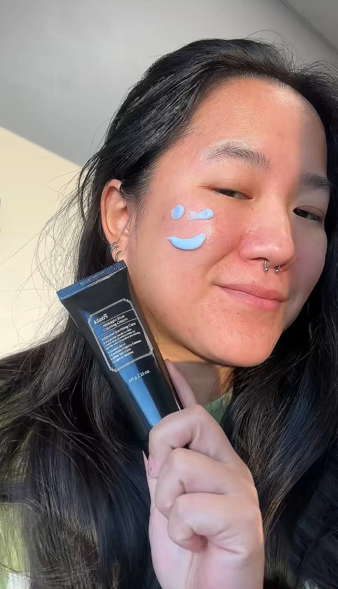 A woman with dark hair holding a tube of Klairs moisturizer, smiling with blue cream on her cheek in a smiley face shape.
