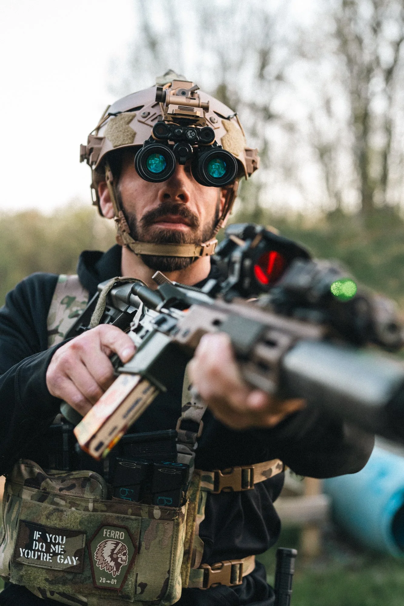 Person in tactical gear aiming a rifle outdoors.