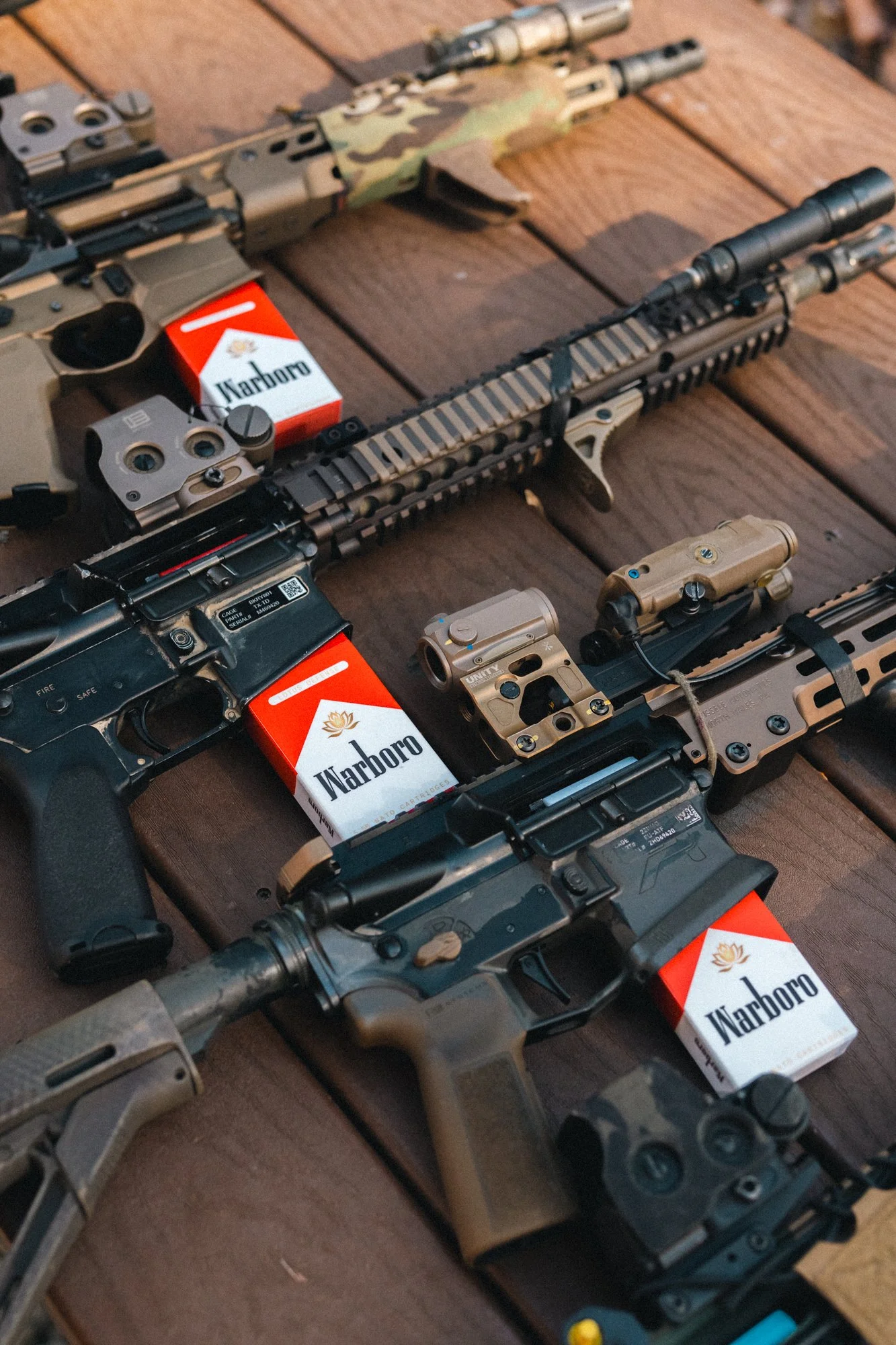 Three rifles with scopes and attachments on a wooden surface, with packs of Marlboro cigarettes placed next to each gun.