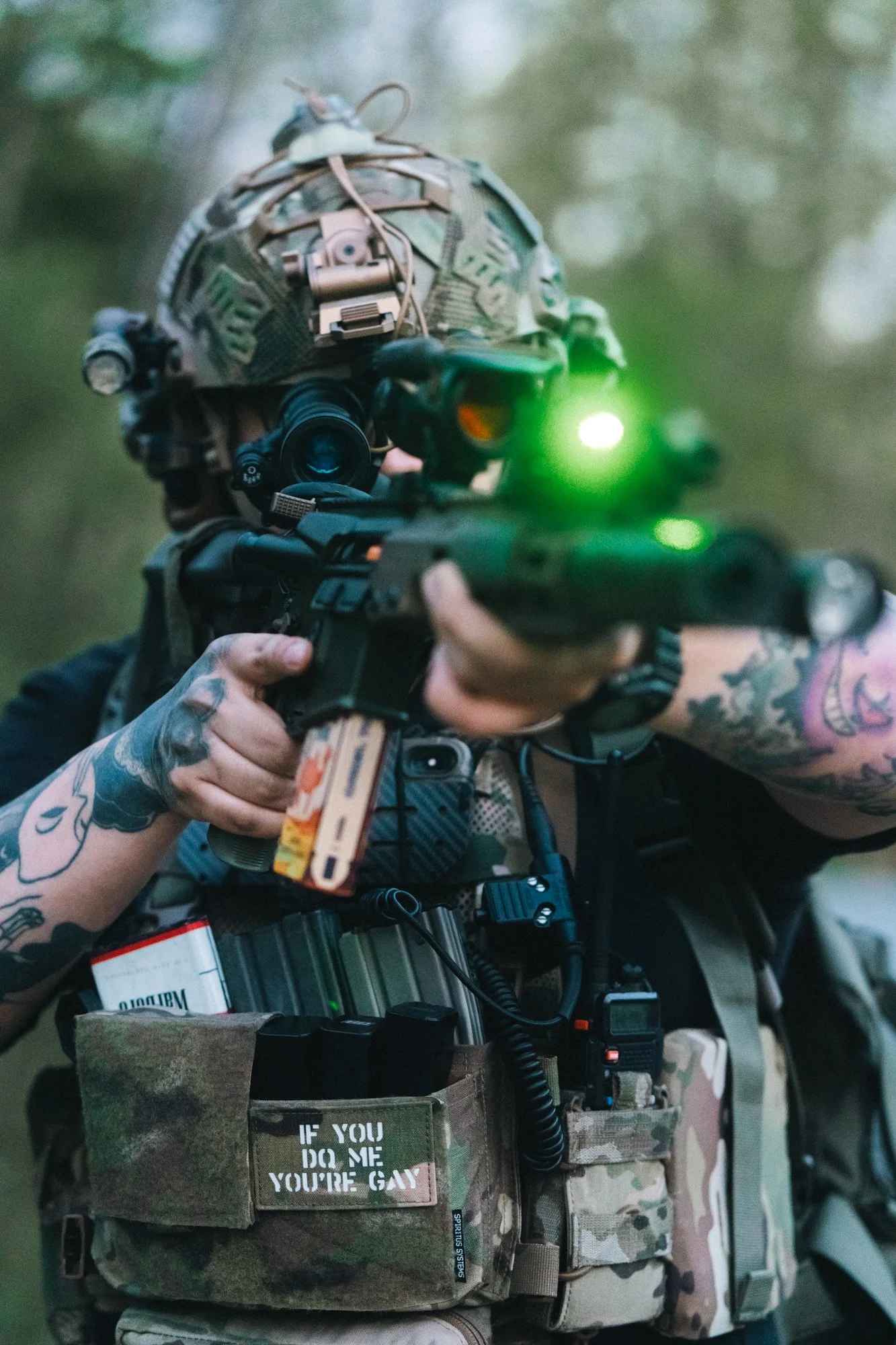 Person in tactical gear aiming a rifle with a green laser, wearing a helmet with attachments, and a vest with patches and pouches.