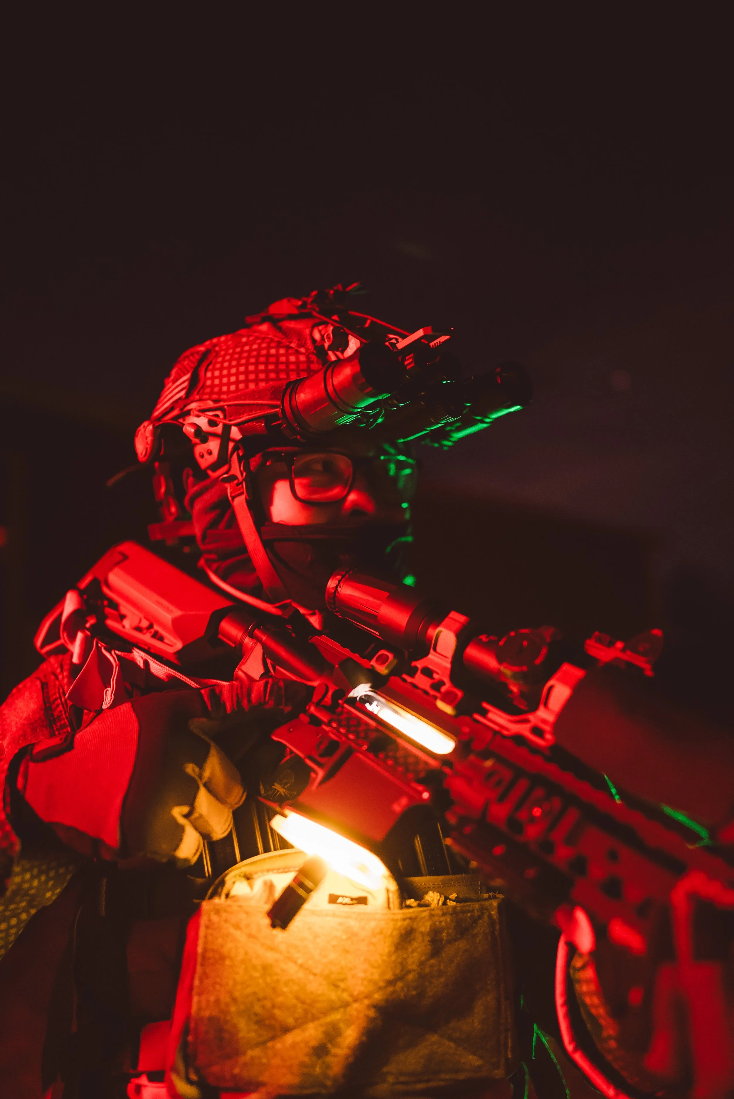 Person in tactical gear with night vision goggles and rifle in dim red lighting.