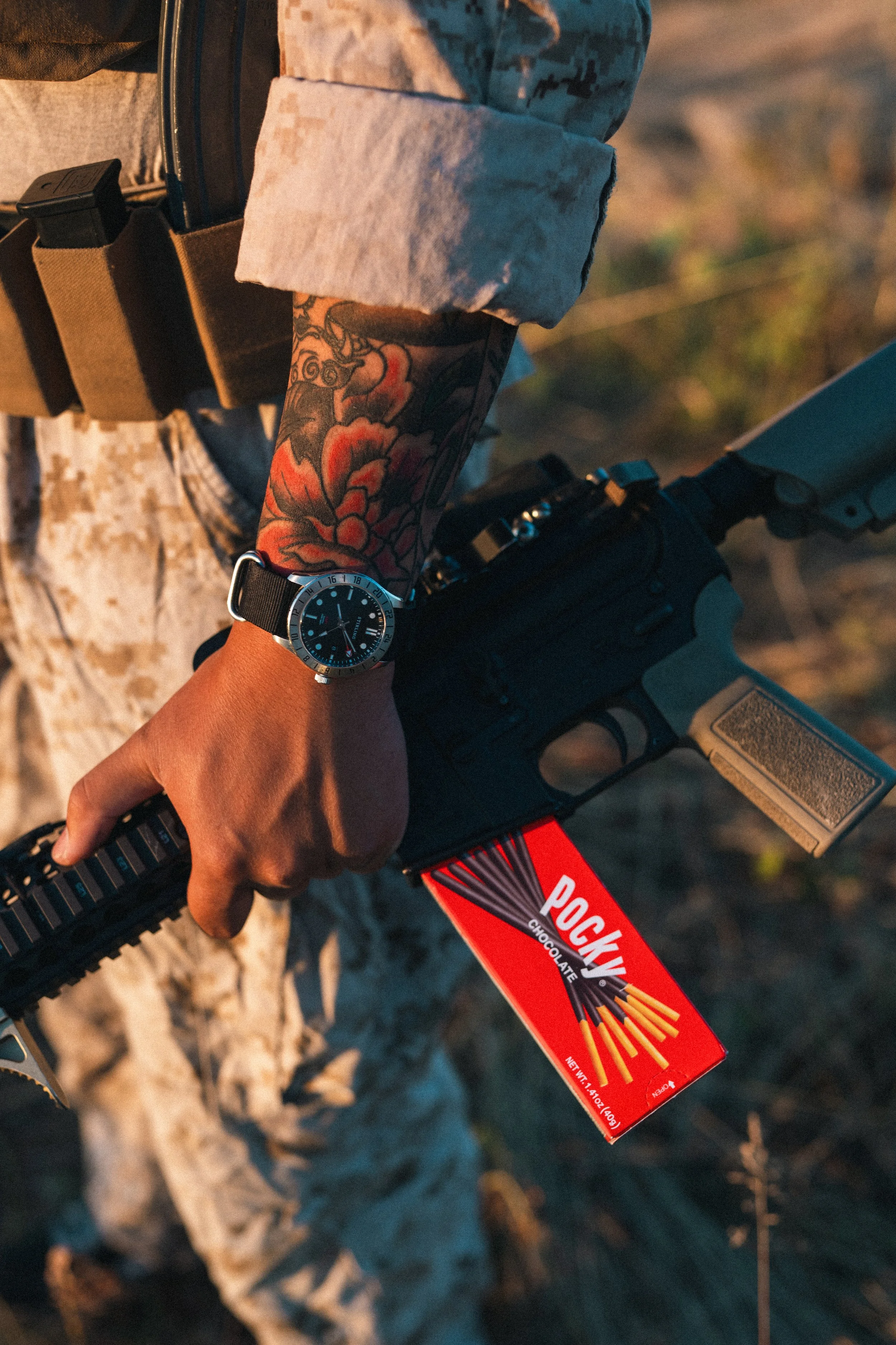 Person in camouflage holding a rifle and a red box of Pocky, with a tattooed arm and wearing a watch.