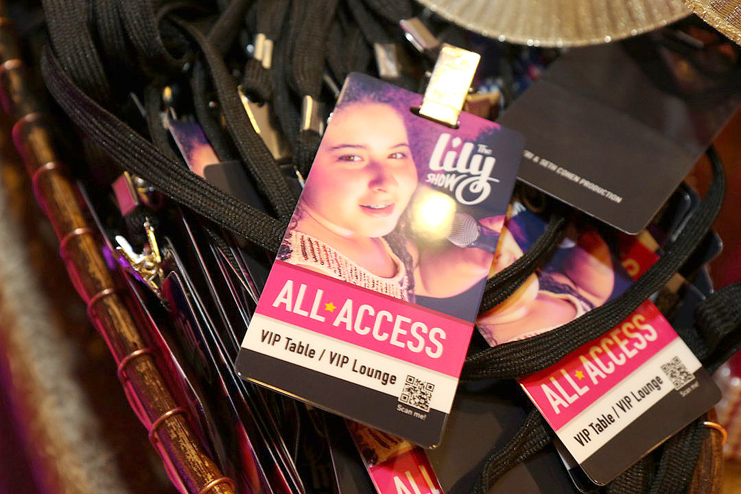Close-up of multiple “All Access” event badges with black lanyards displayed in a basket. Each badge features a photo of Lily holding a microphone and singing, with white text over pink, white, and black background elements.