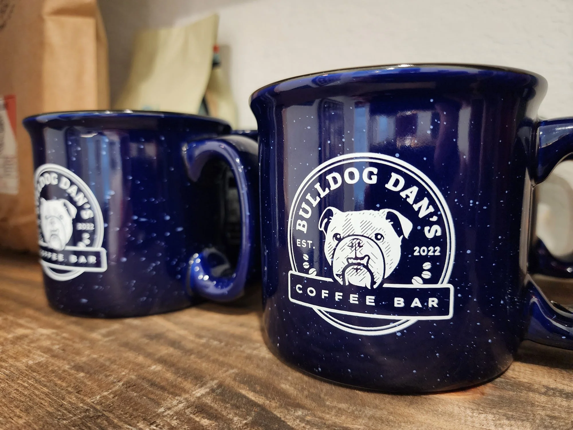 Close-up of two dark blue mugs with small light-blue speckles featuring the white Bulldog Dan’s Coffee Bar logo, displayed on a wooden shelf beside brown paper bags of coffee beans.