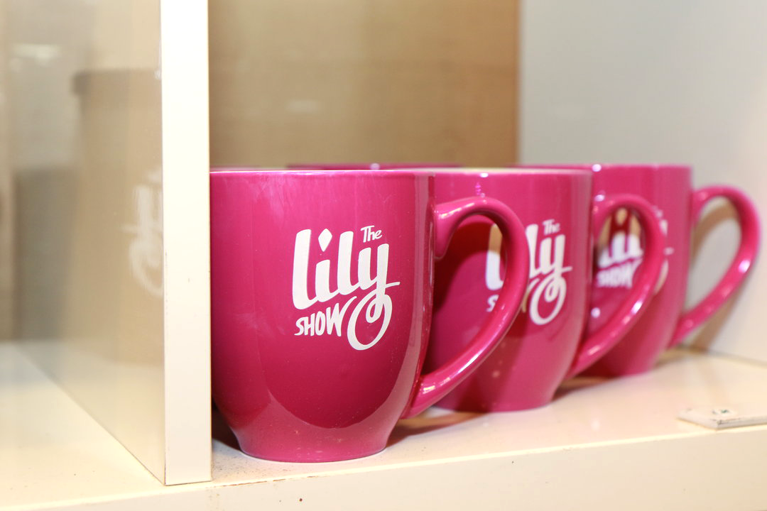 Close-up of three pink coffee mugs featuring The Lily Show logo, arranged uniformly side by side in a cubby as part of a merchandise display.
