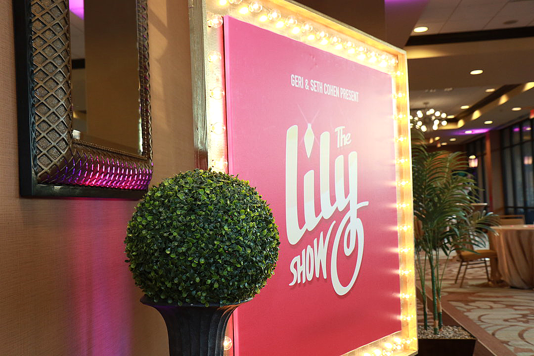 Large gold marquee sign with lights featuring The Lily Show logo in white on a pink background, displayed in a hotel hallway outside a ballroom with plants and event tables in the background.